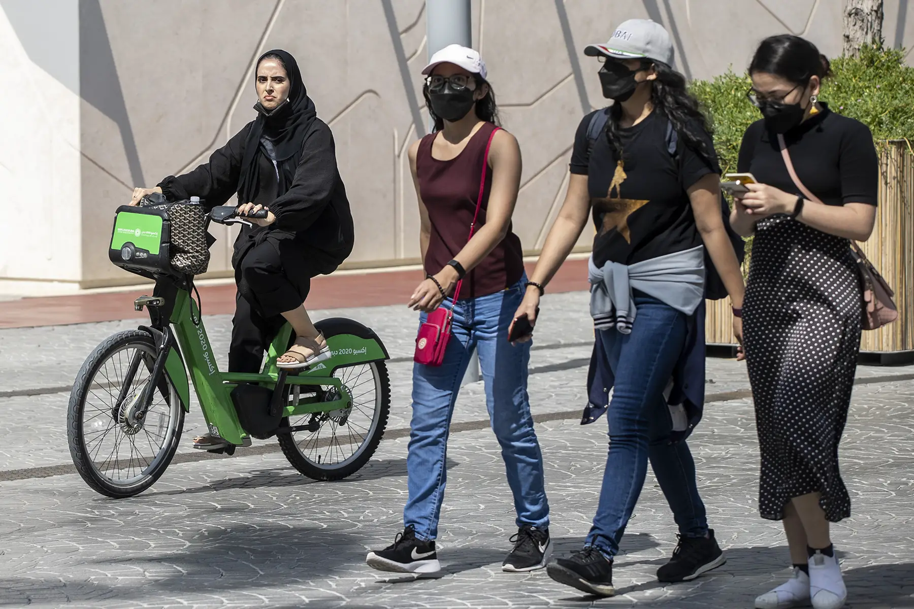 Three women walking wearing t-shirts, jeans, skirts. One woman on a bike in a headscarf.