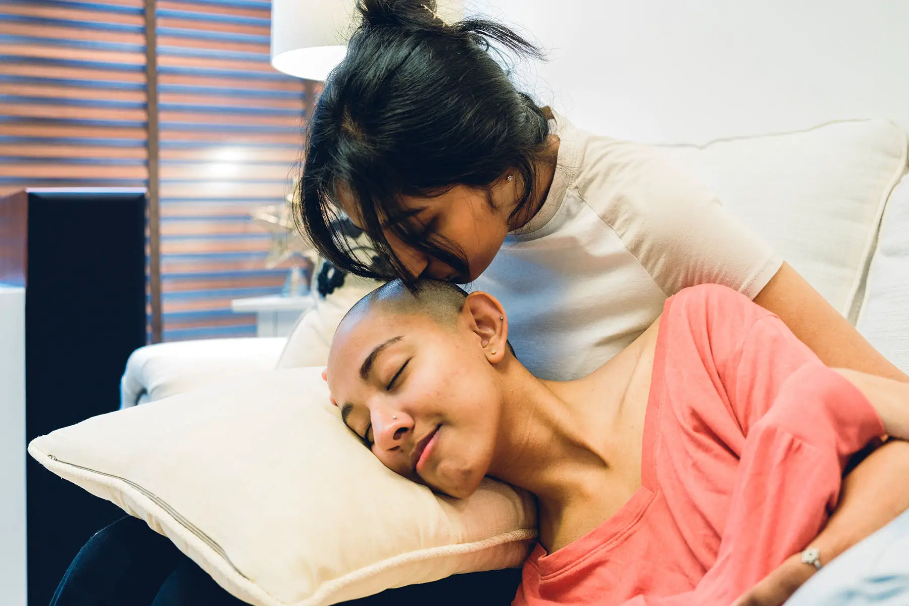 Girl with cancer lying on a pillow on someone's lap, who is leaning over to kiss her head.