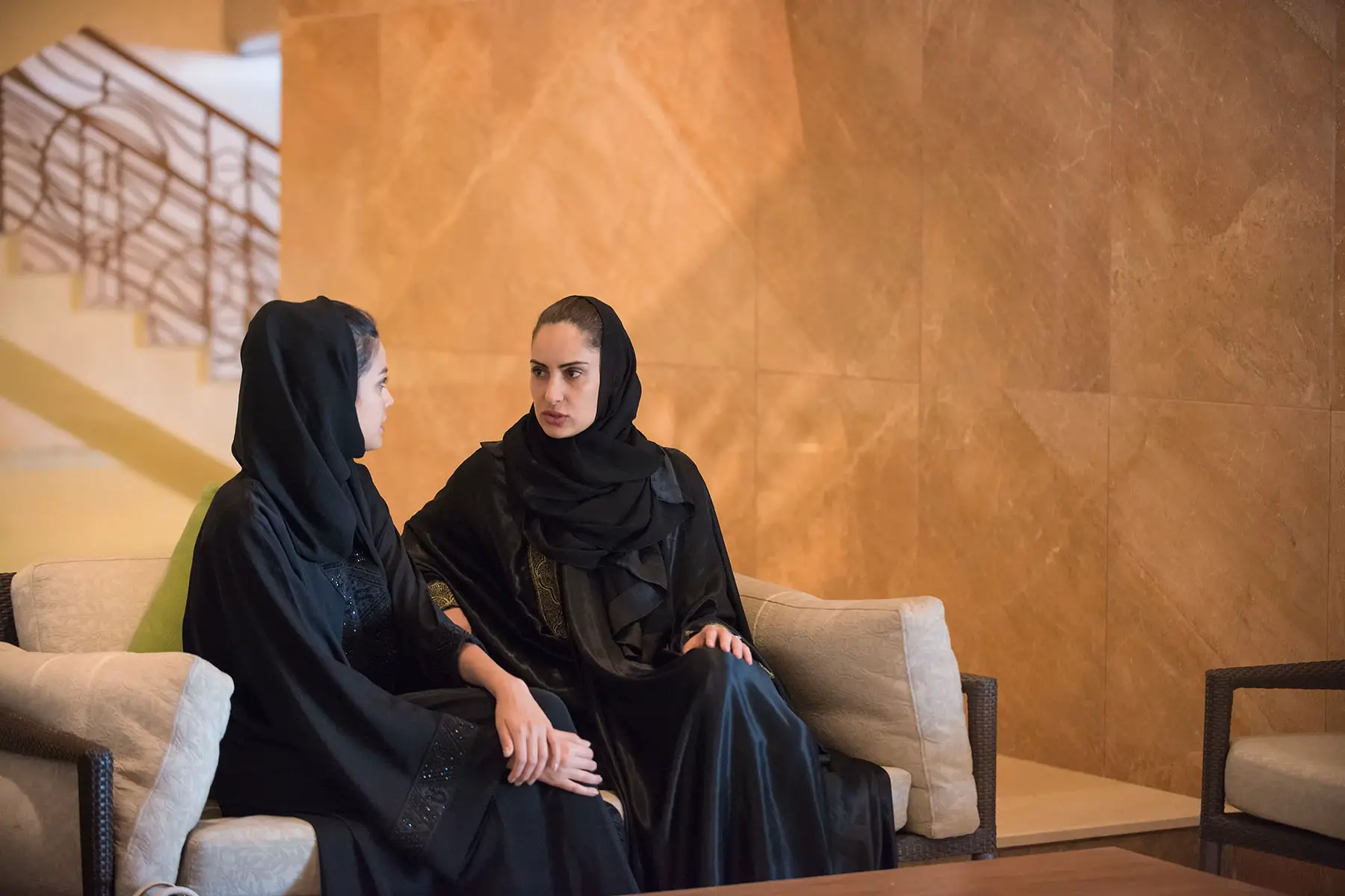 Two women wearing a traditional black abaya and hijab have a conversation in the lobby of a luxury spa resort hotel.