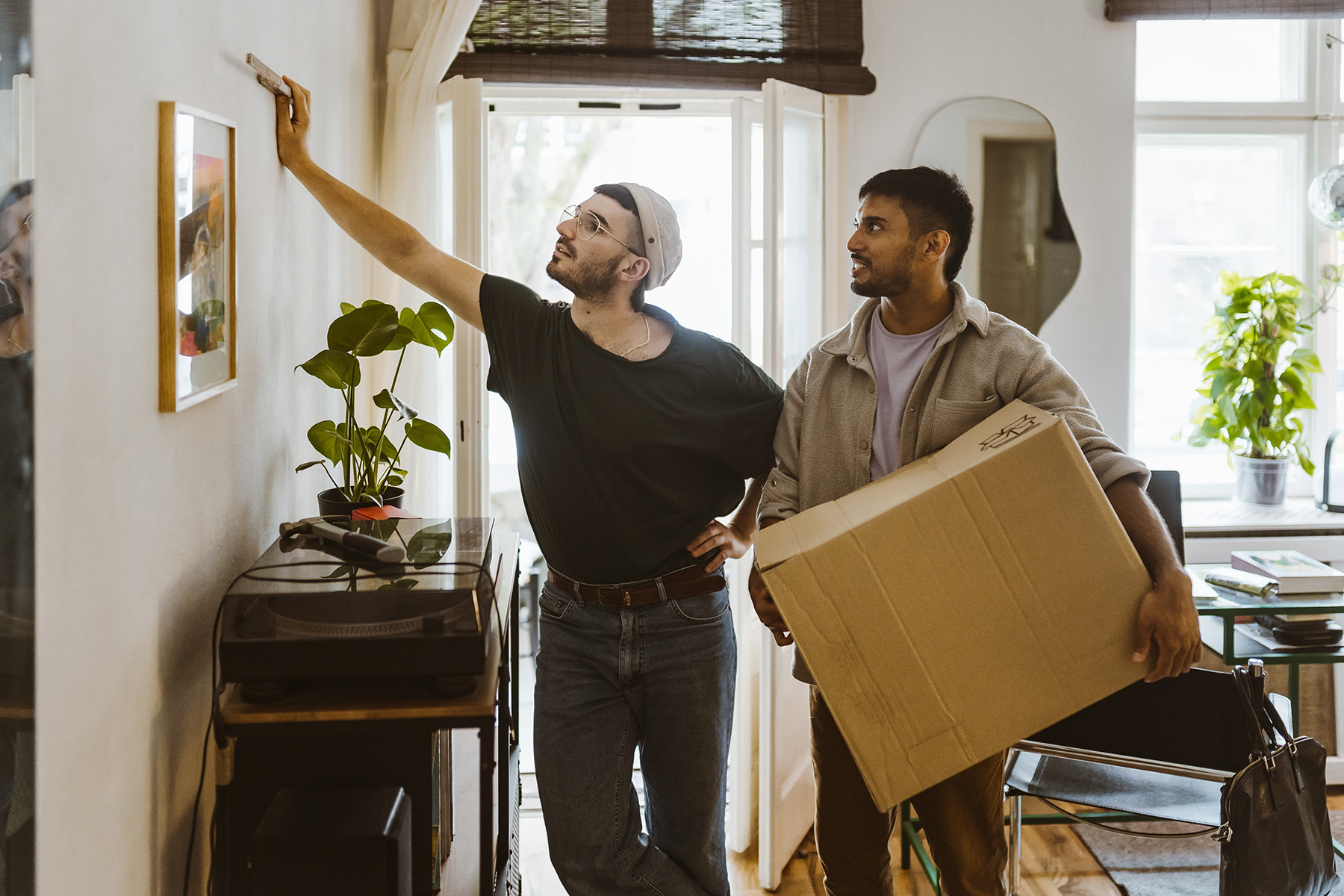 Two guys - one holding a moving box - walk into a home - one measures something on the wall