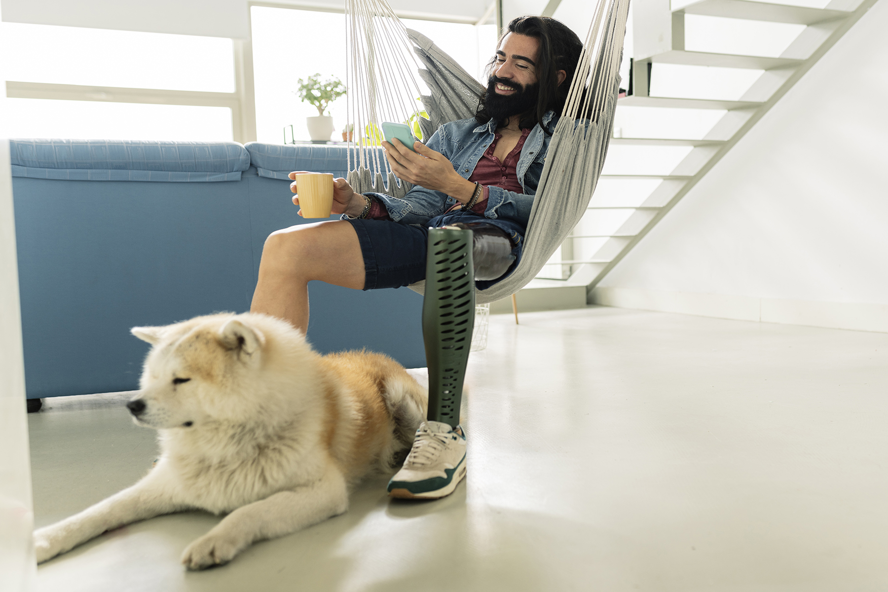A man sits in a hangmat inside his home with his dog, smiling at his phone. He has a prosthetic leg.