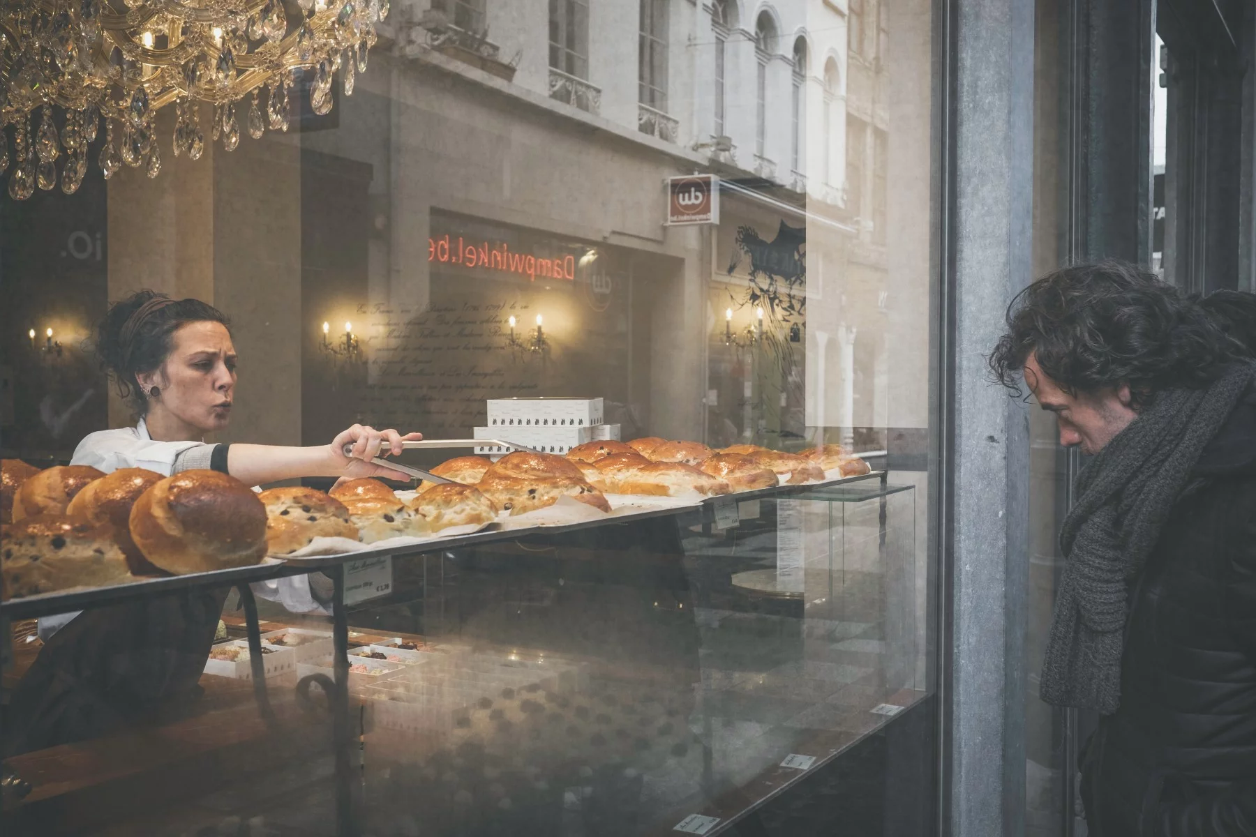 Woman working at a bakery looking angry while she is grabbing one of the buns in the window display. Man is looking inside the shop.
