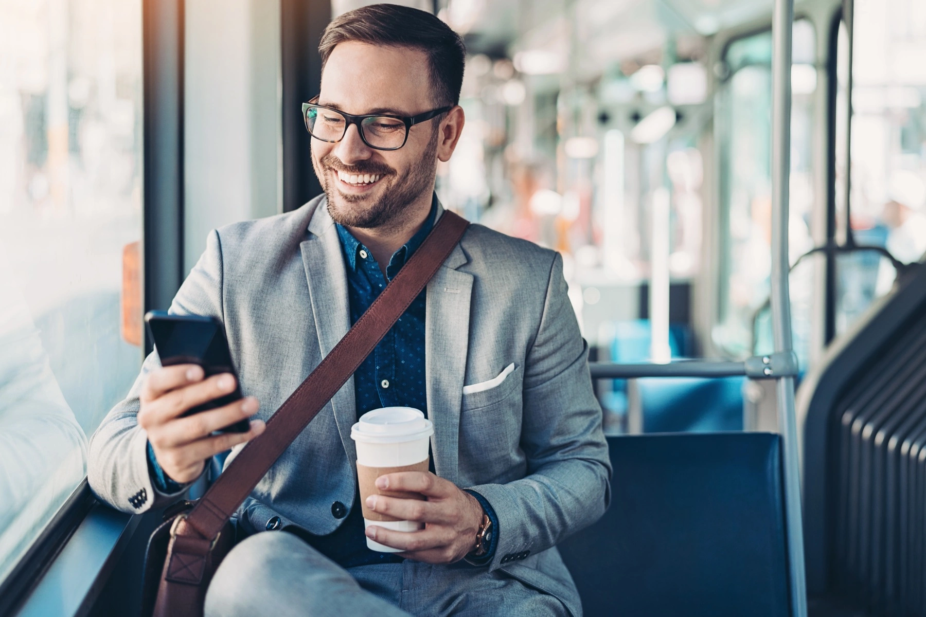 a businessman wearing a suit and sitting on a bus smiling as he holds his mobile and a cup of coffee