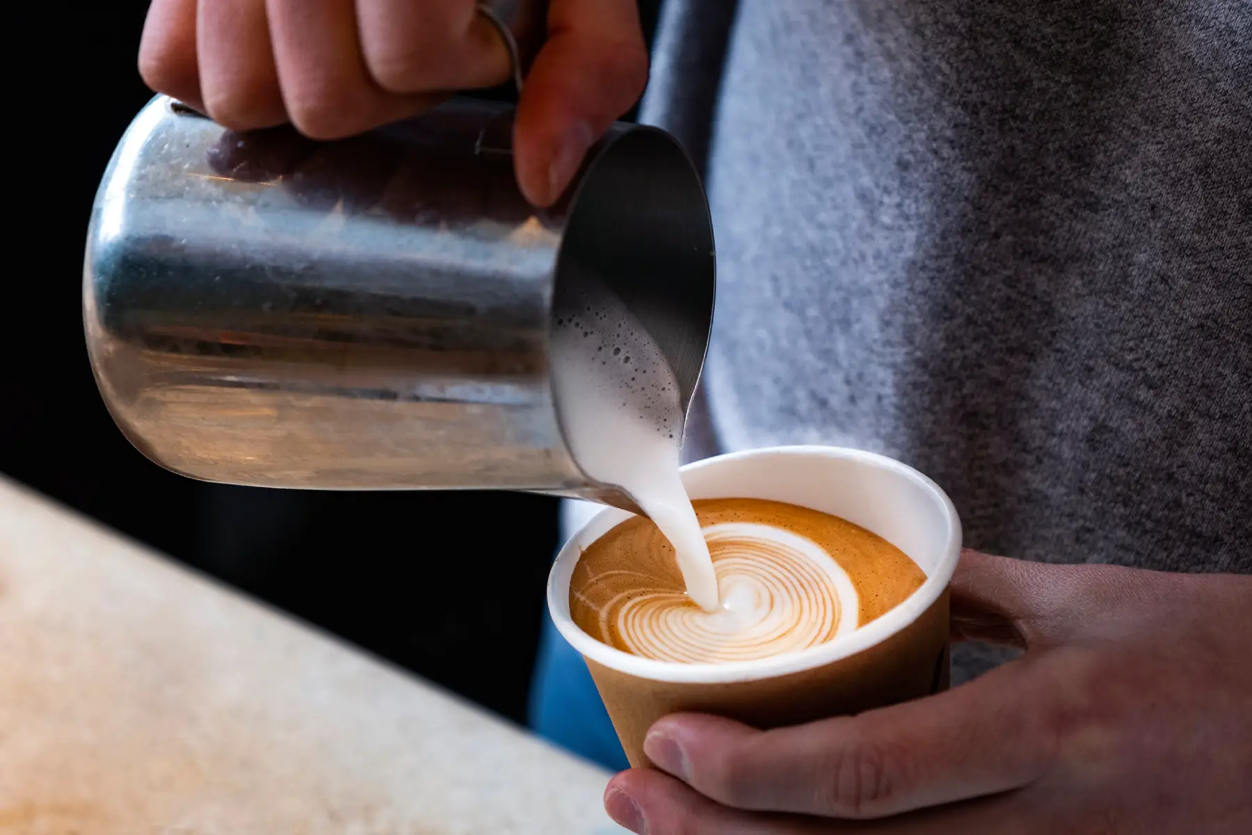 a man's hands holding a cup of fresh coffee and pouring milk into it to make pretty latte art at a coffee shop