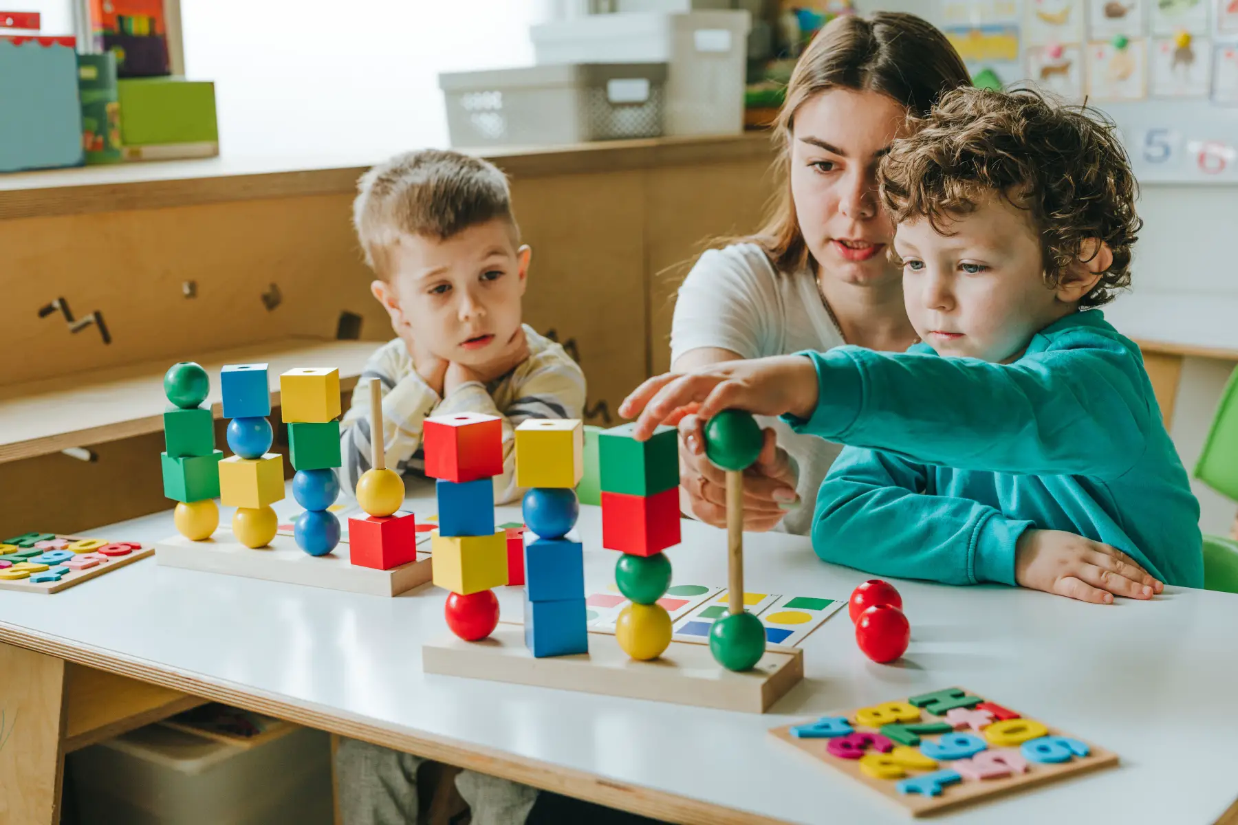 a female teacher helping a little boy sort different colored wooden cubes on his desk in a preschool classroom setting