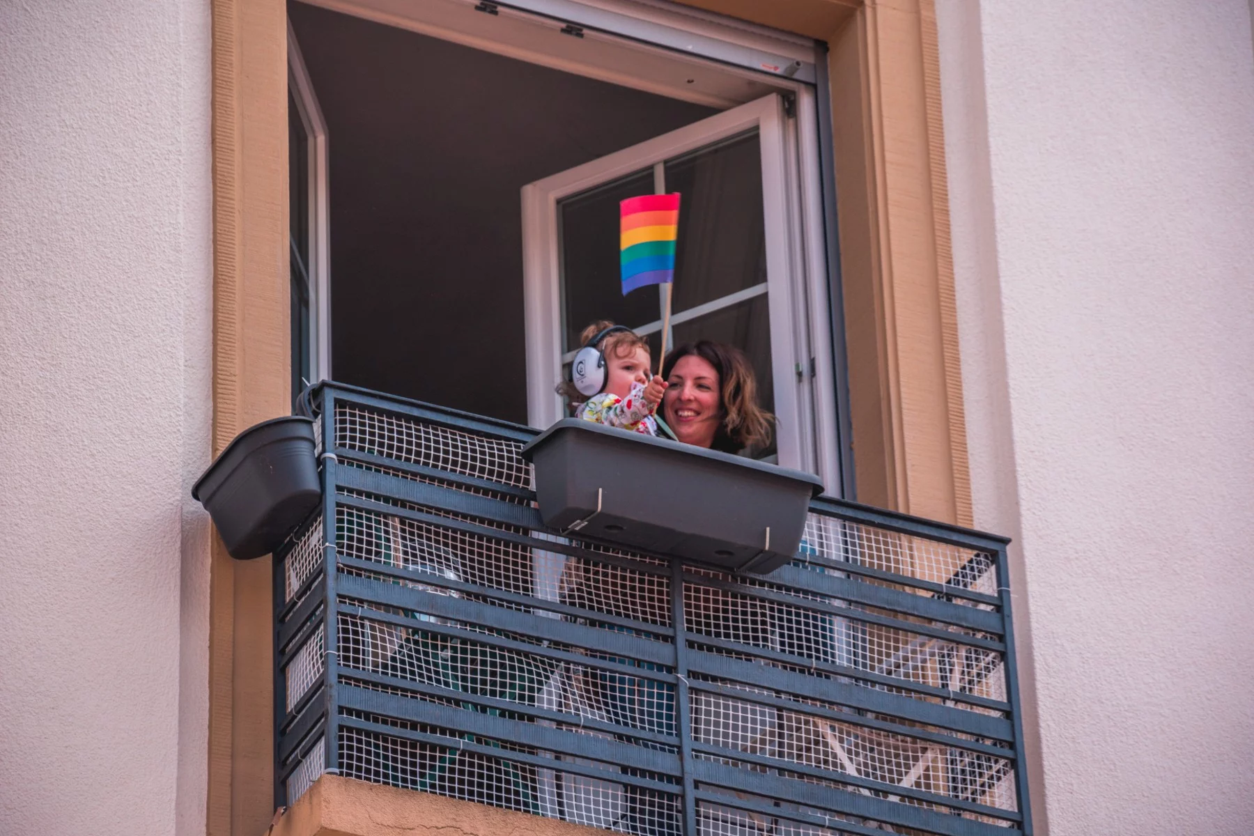Mother and baby standing at a balcony. The baby is holding a rainbow flag, while their mother looks at them adoringly.