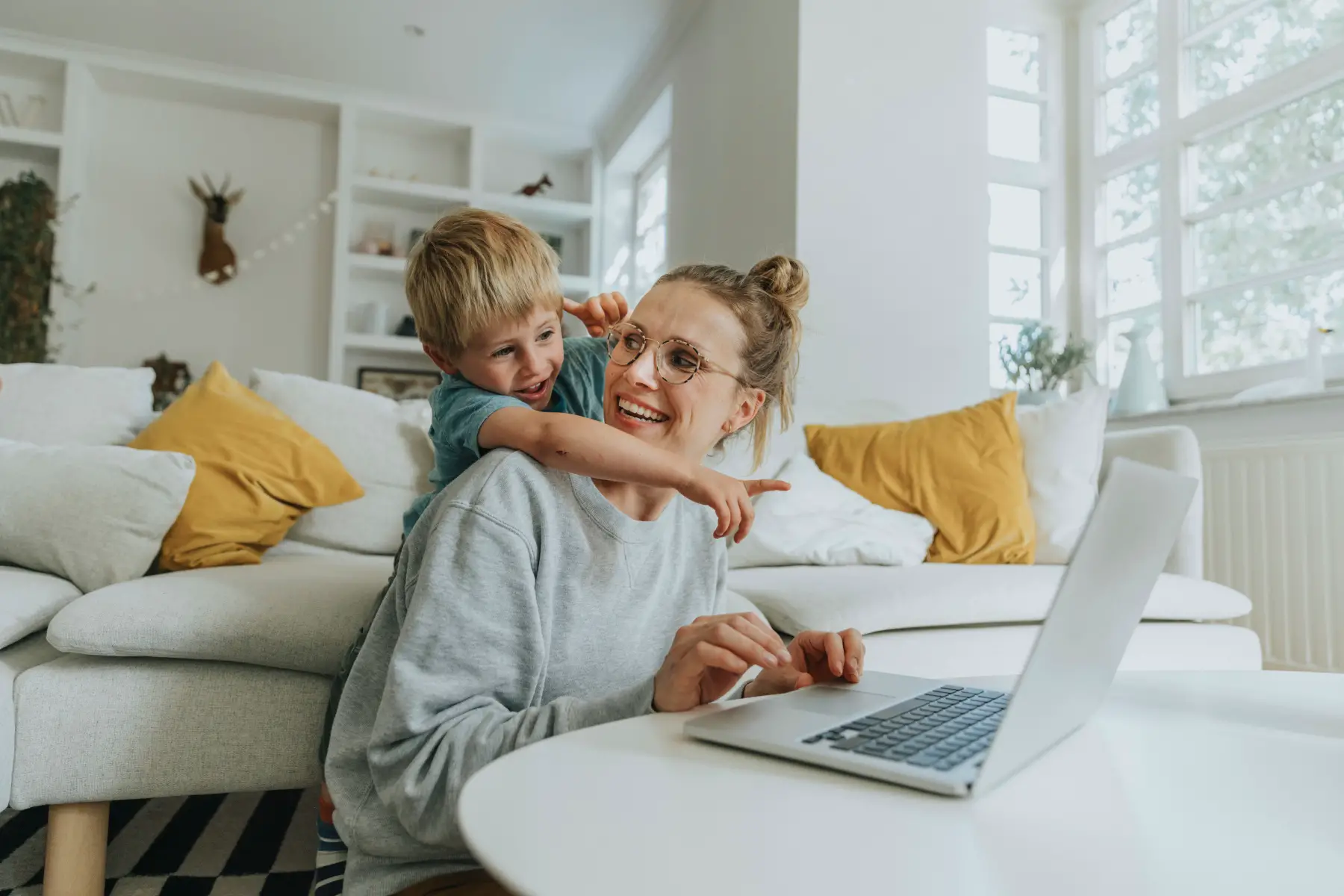 a mother sitting on the floor in her lounge with her laptop as her young son looks over her shoulder