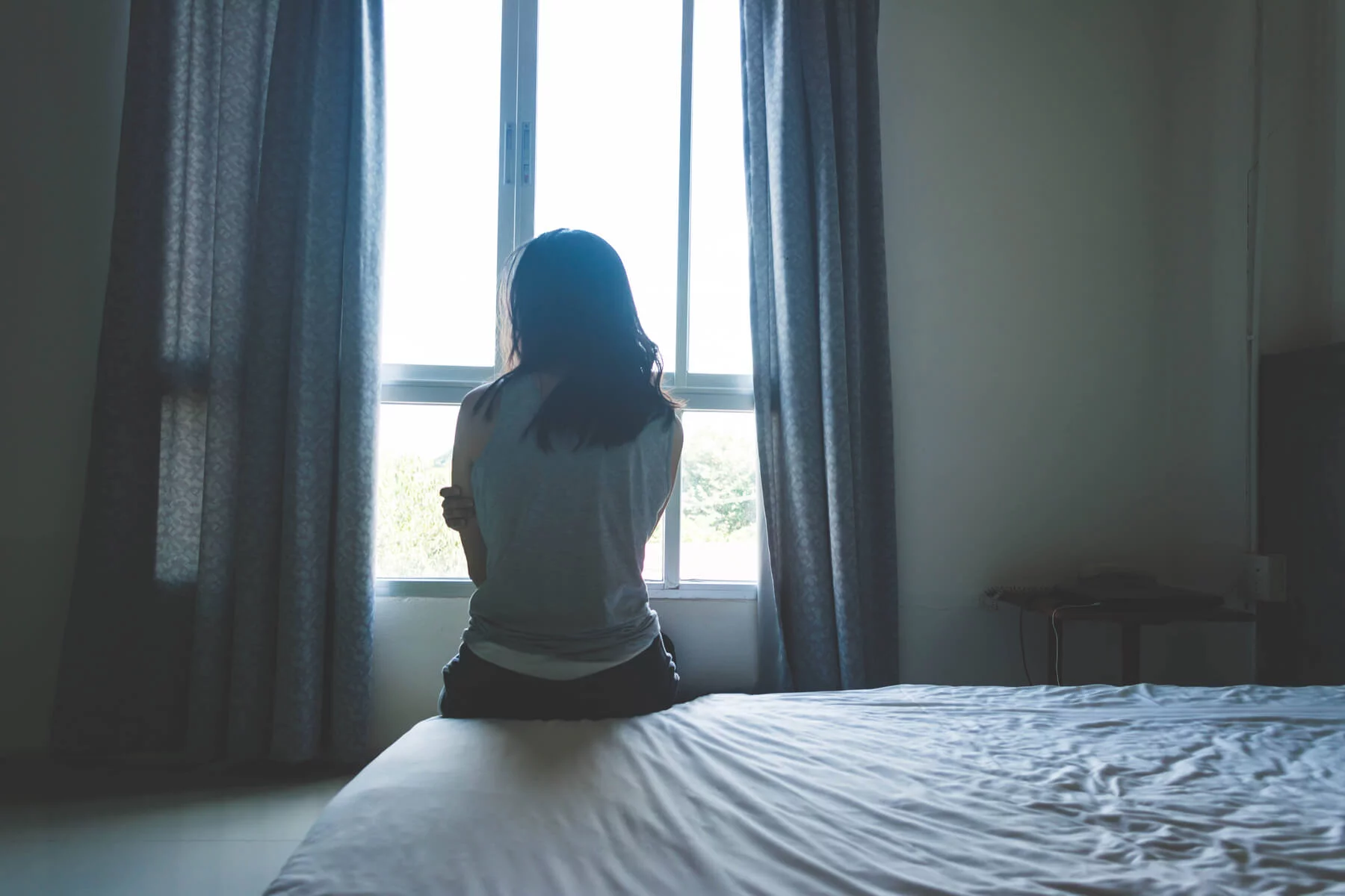 a woman sitting alone on a bed facing the window