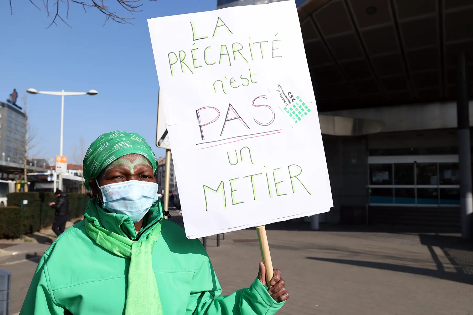 A woman in a green jacket and surgical mask protesting with a sign that says 'La précarité n'est pas un métier' - 'precarity isn't a job'