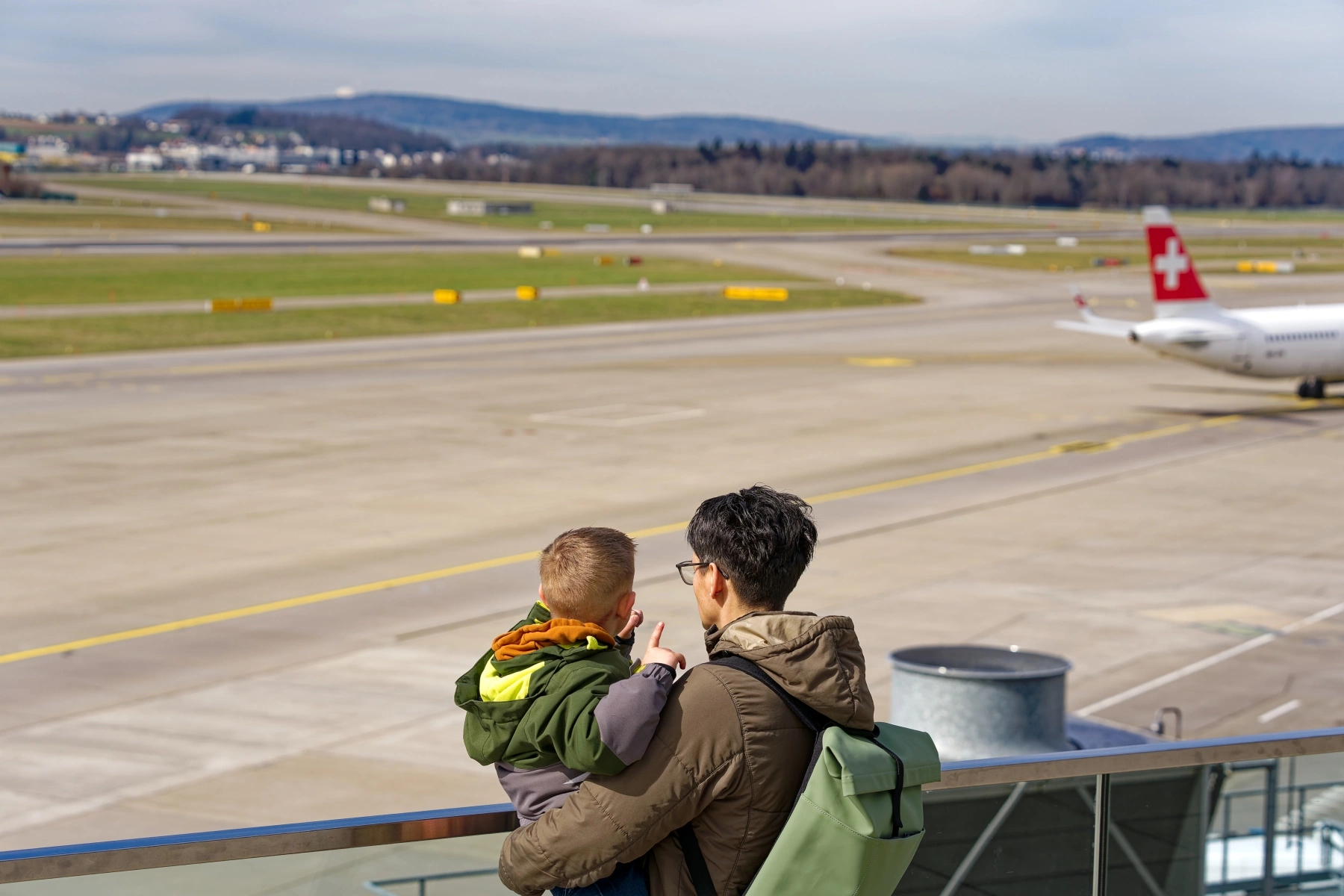 Father holds child as they look out at an airplane runway with s Swiss plane