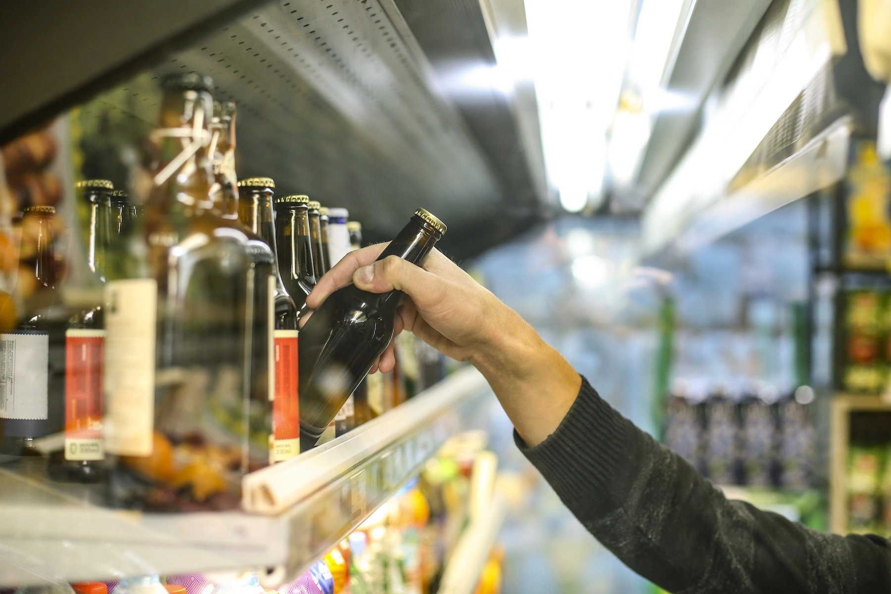 Close-up of a man's arm as he reaches for a single bottle of beer on a refrigerated store shelf