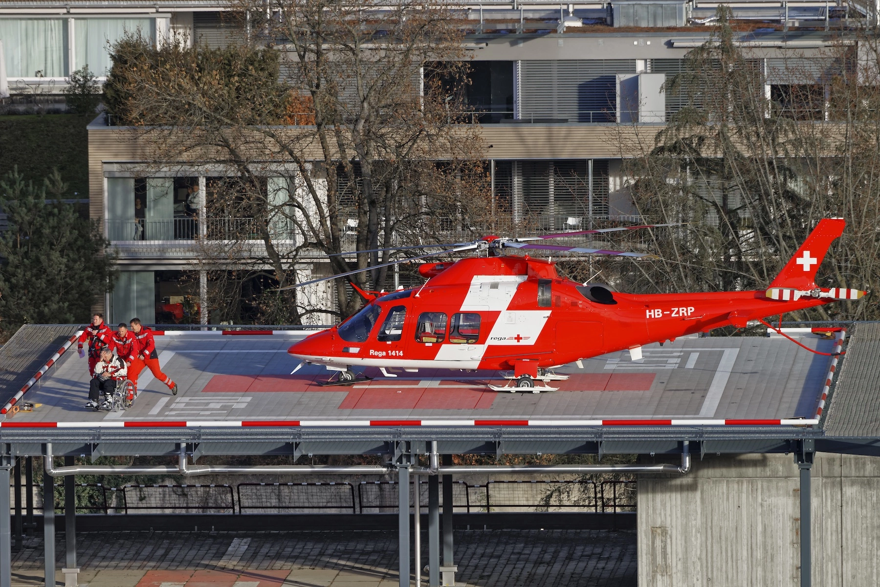 A helicopter sits on the helipad of a hospital in Thun, Switzerland while medics transport a patient in a wheelchair