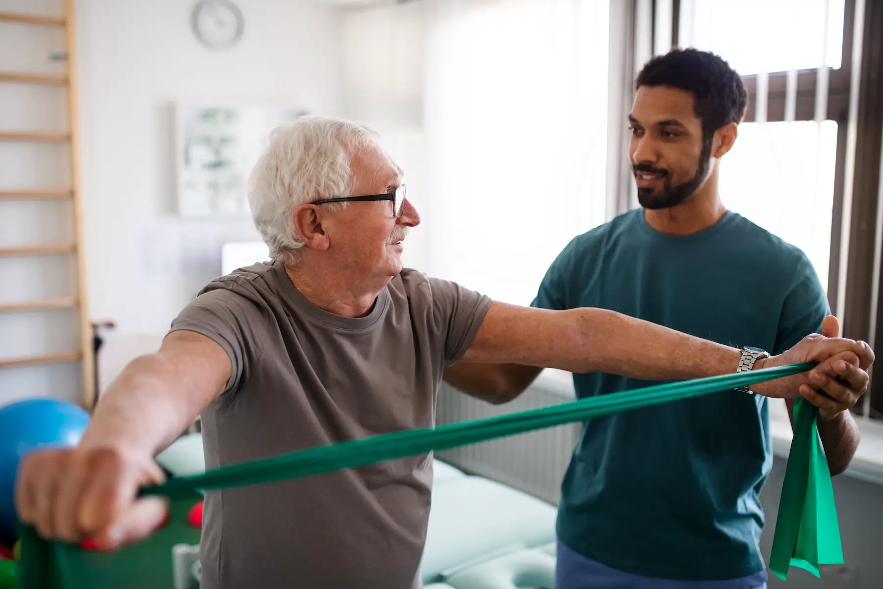 An older man holds a stretch band in both hands, working on his strength, supported by a younger male physical therapist