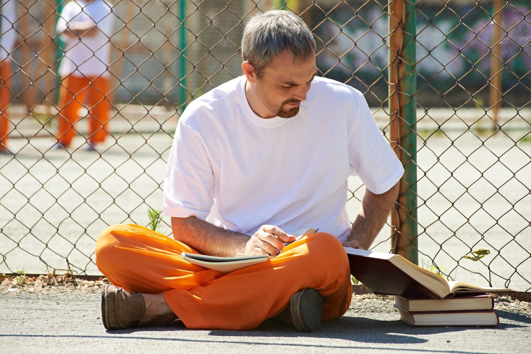 A man in prison attire studies with books and a notebook in a prison yard during the daytime
