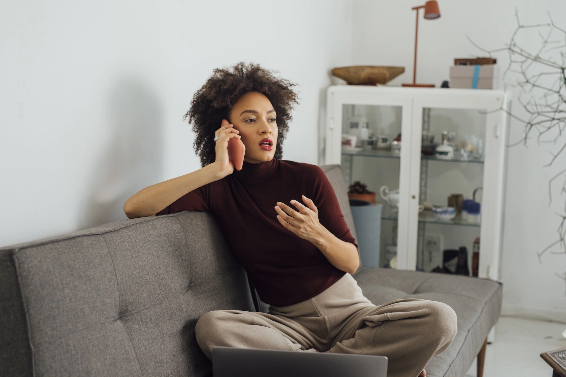 A woman sits speaking on her smartphone in her living room, with a serious expression