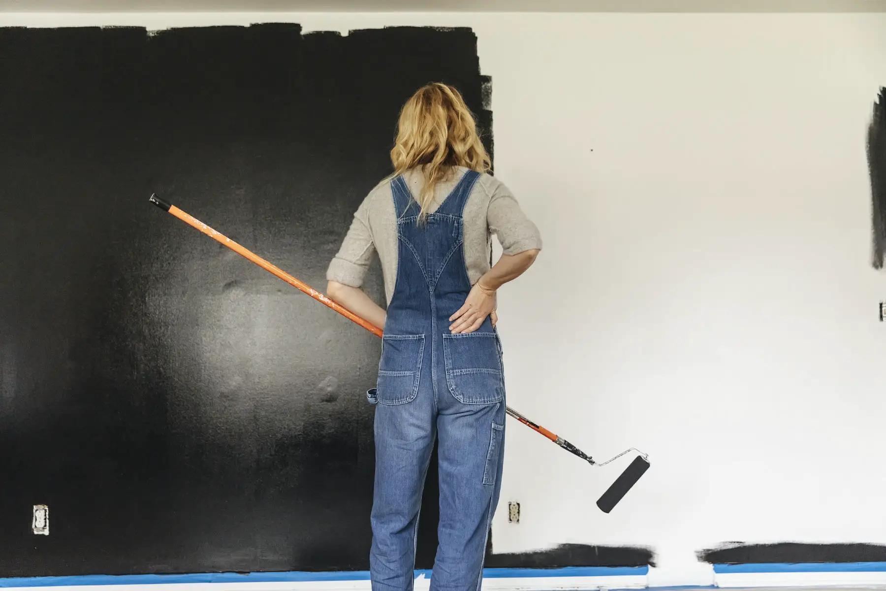 Woman in overalls is looking at her work, halfway through painting a wall entirely black.