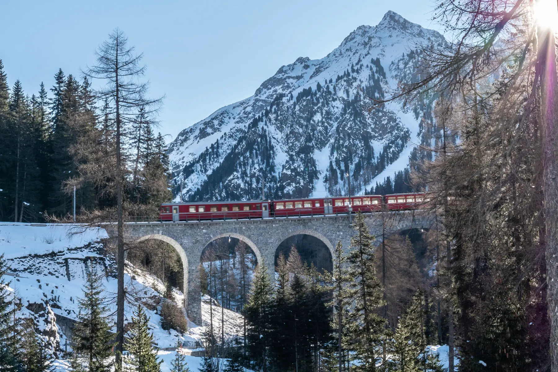 Red train on old viaduct in Switzerland, sunny winter morning in Graubunden Canton. Snow and sunbeam