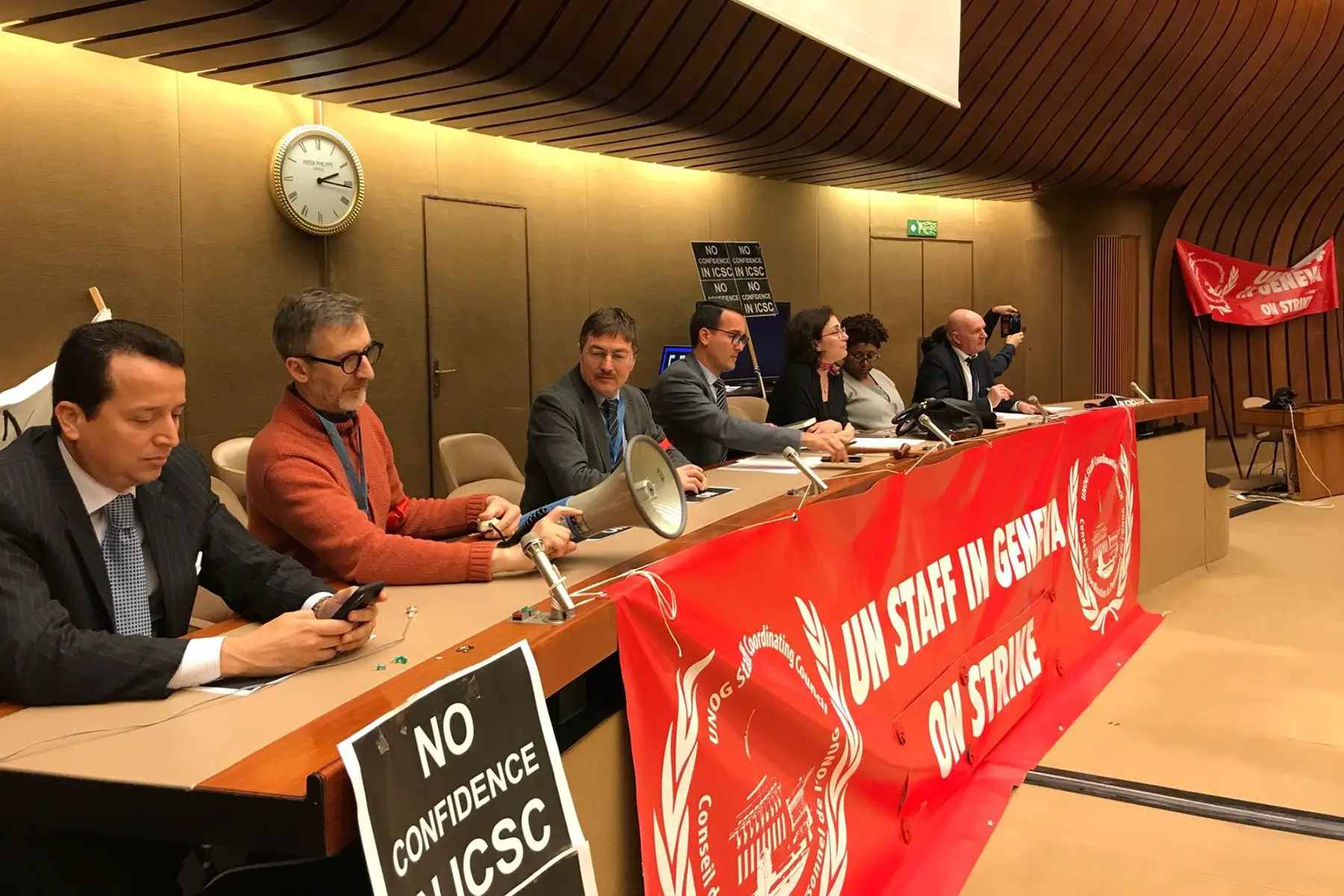 Staff at the UN on strike, sitting at a desk with a large red banner reading 'UN STAFF IN GENEVA ON STRIKE'
