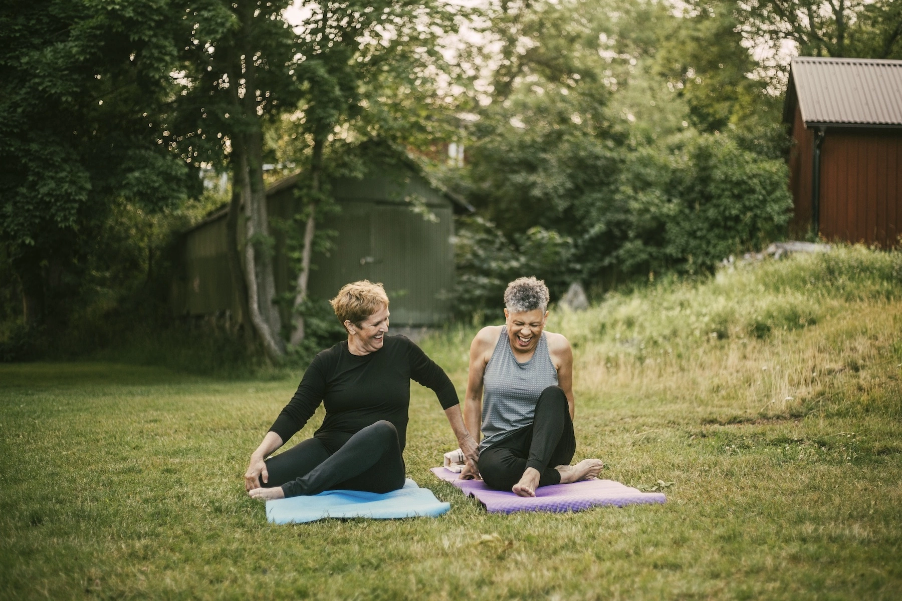 Two older women sit on yoga mats in the grass in the backyard, they are laughing and holding hands