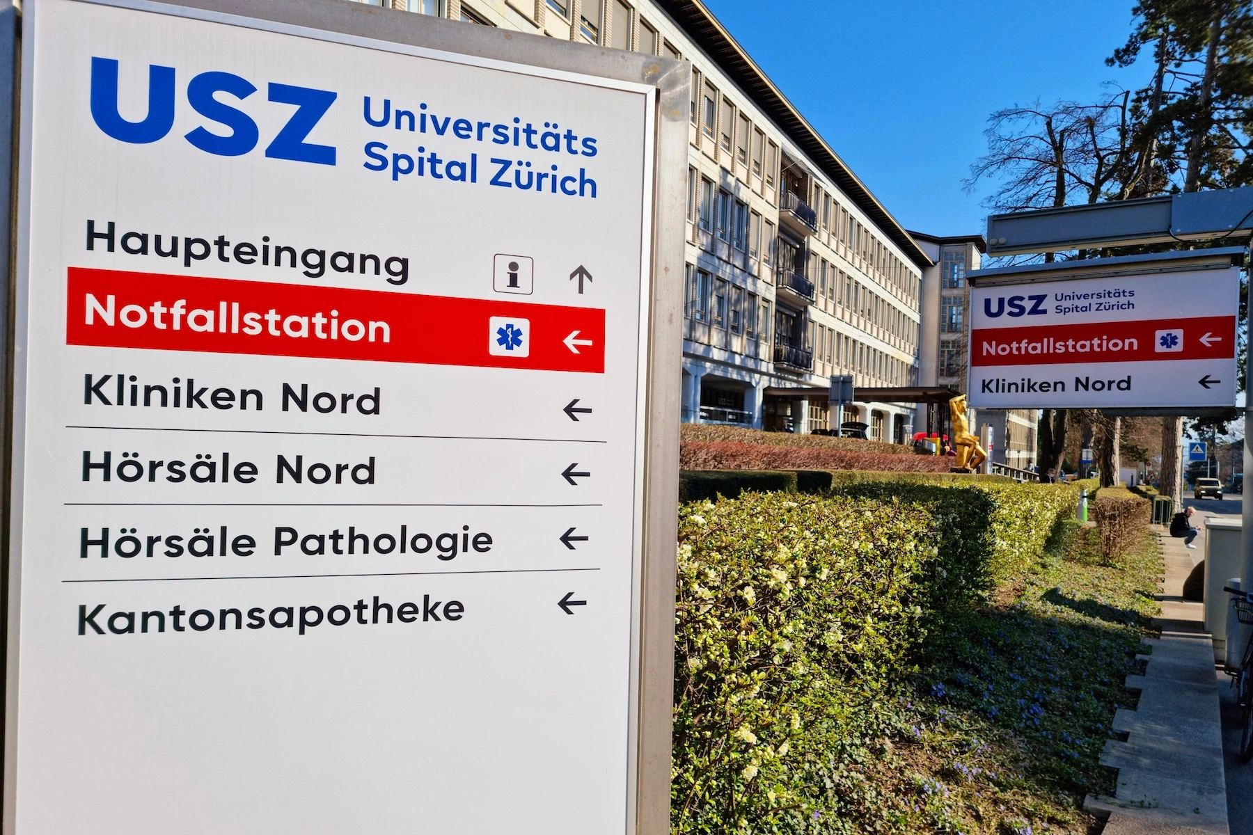 The University Hospital of Zurich displays a sign outside showing the way to the entrance, emergency drop-off, and various departments.