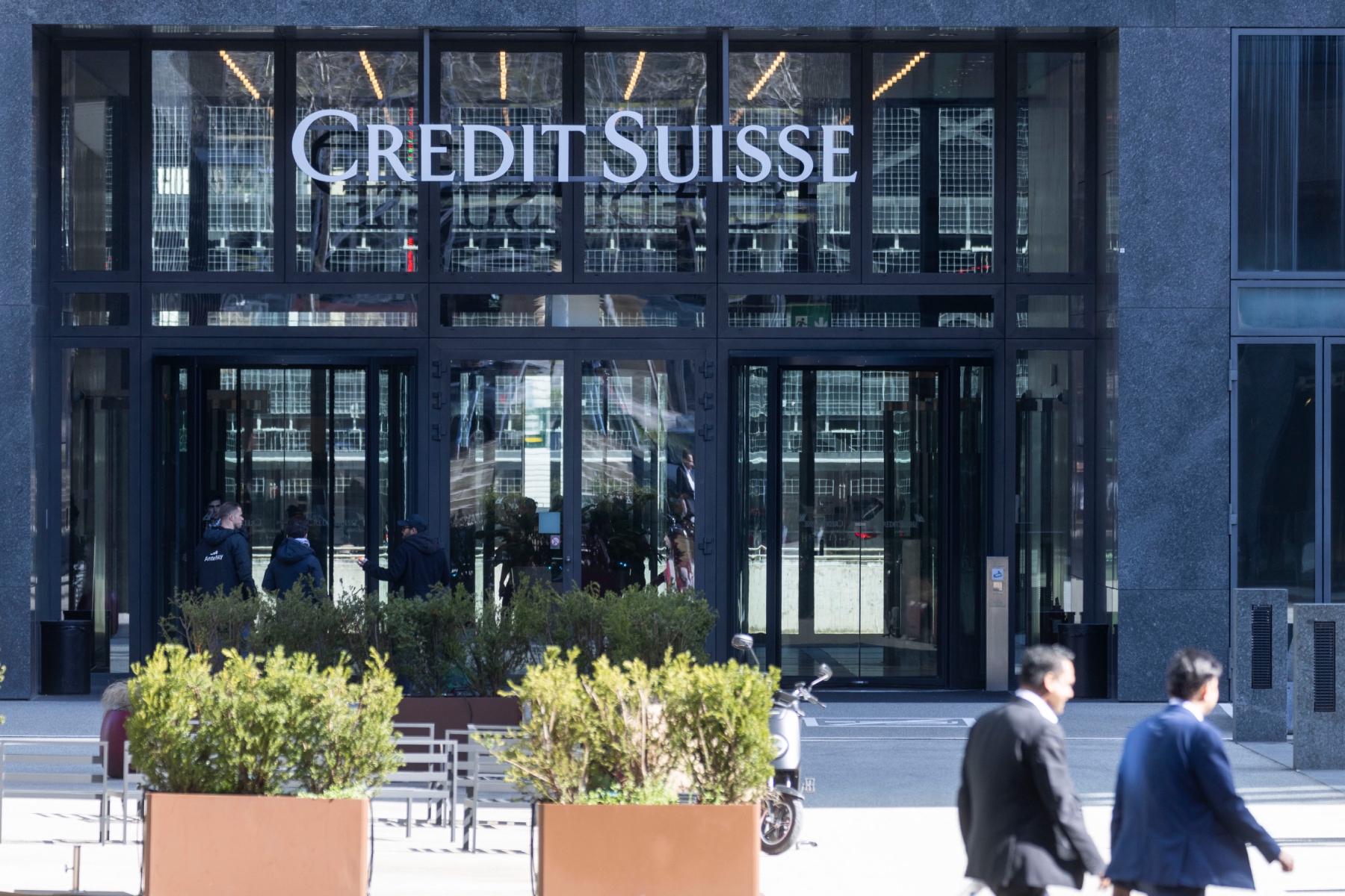 Credit Suisse offices exterior, with two large potted plants outside and two business men walking past 
