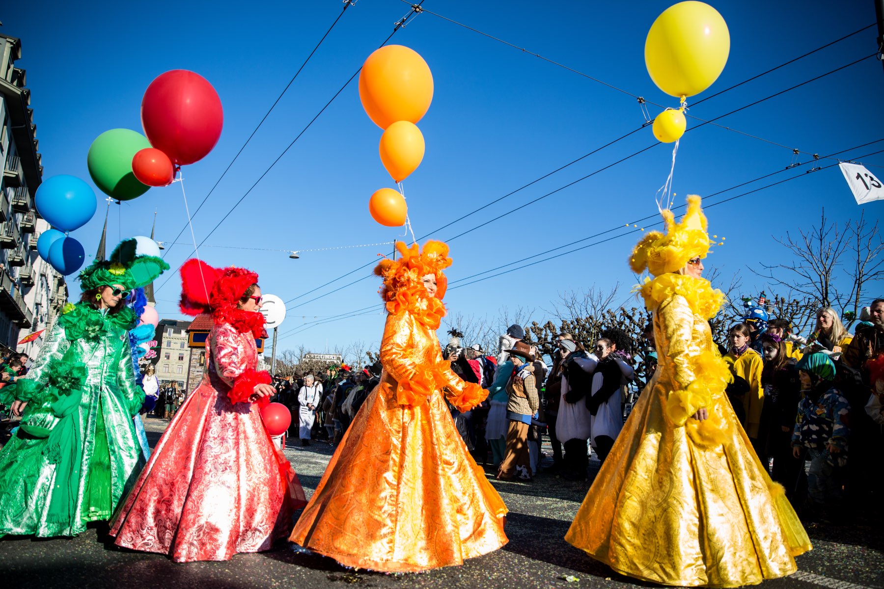 Four women dressed individually in yellow, orange, red, and green ballroom dresses carry balloons as part of Lucerne's carnival celebrations