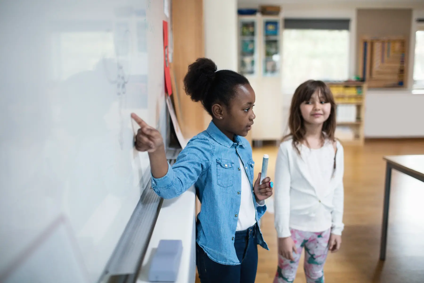 two young girls of different ethnicities (black and caucasian) standing next to a whiteboard in front of the class