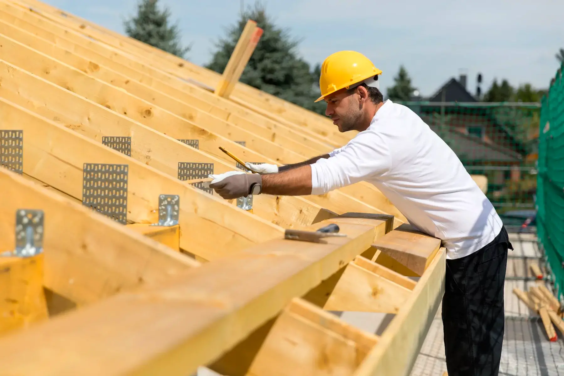 A construction worker wears a safety hat and gloves while focused on building a wooden roof