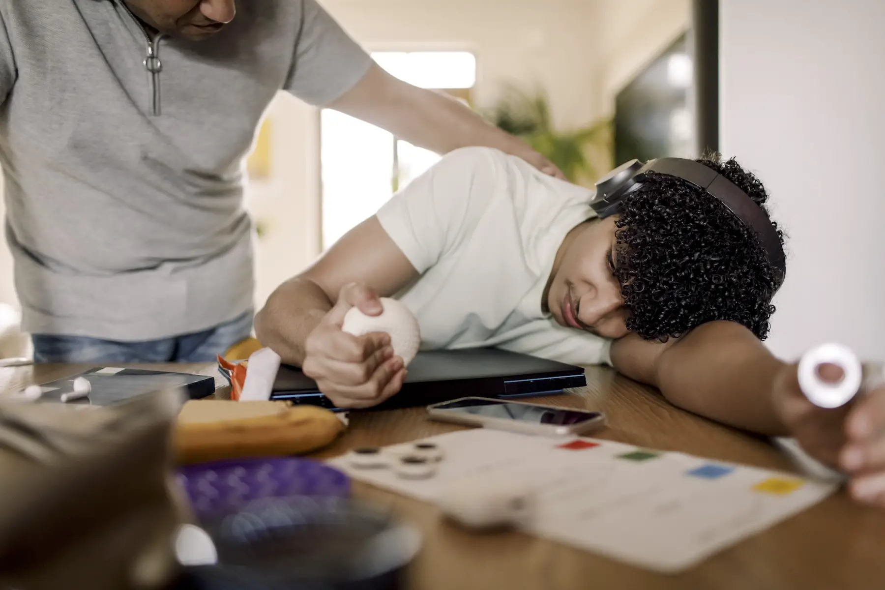 A father comforts his teenage son who is laying his head down on a desk on top of his homework