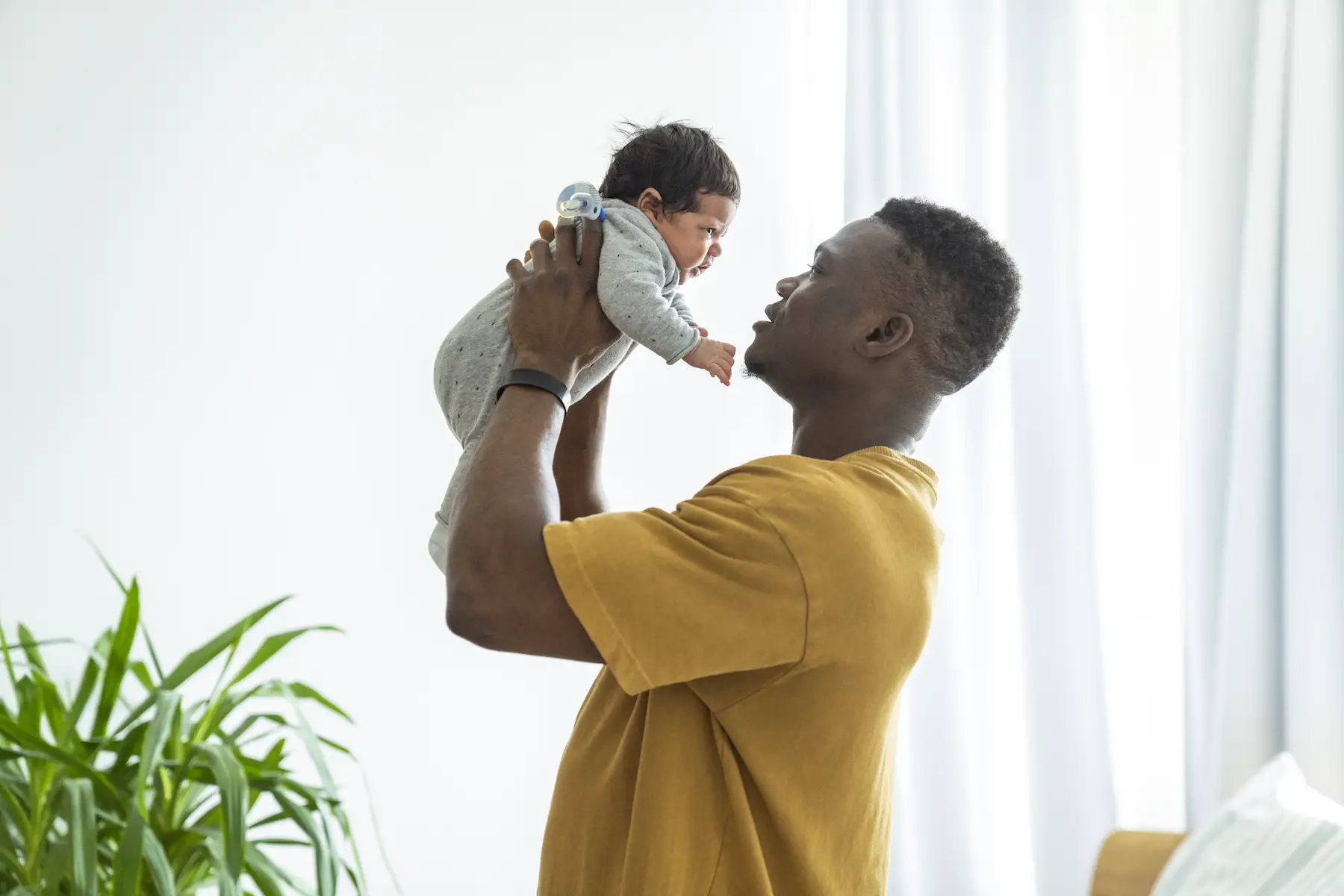 A father plays with his newborn baby boy, lifting him up to eye level in a brightly lit room.