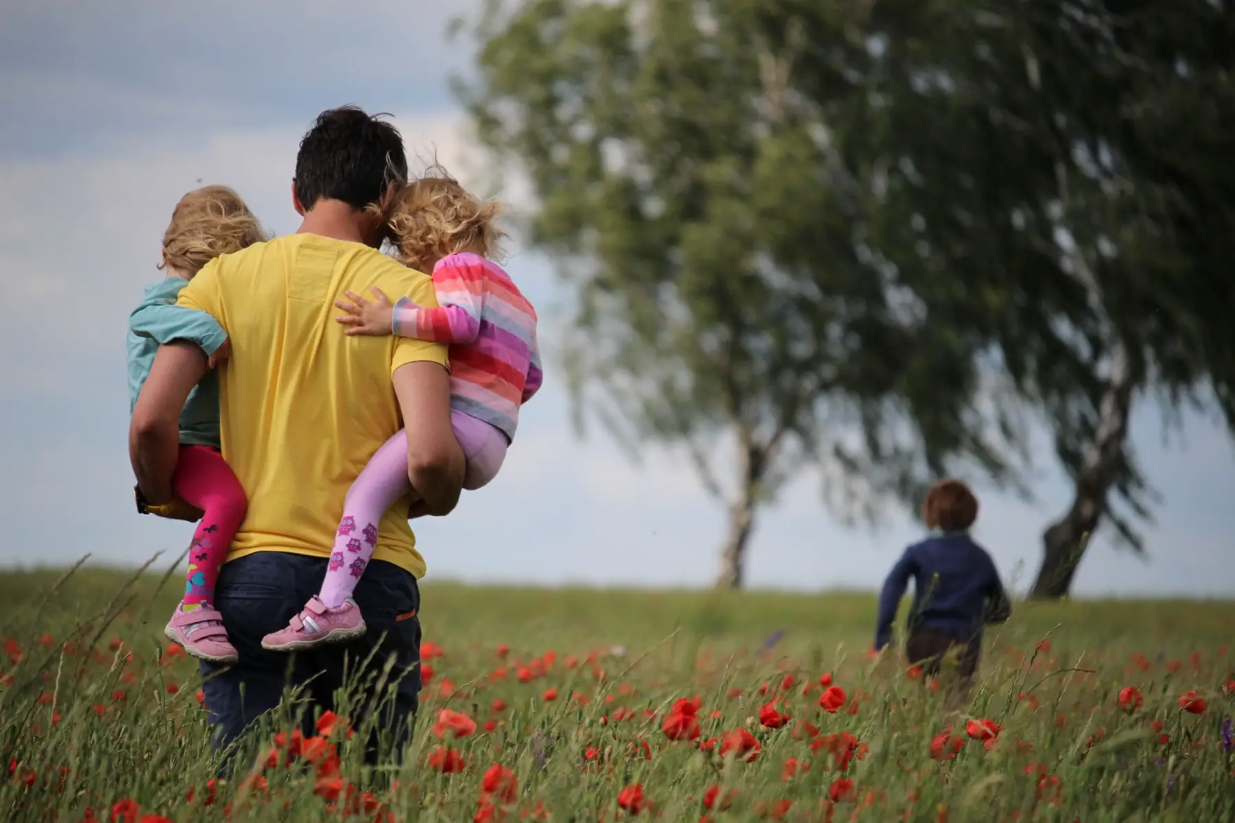 a man carrying his two small children in his arms as he walks across a field, away from the camera