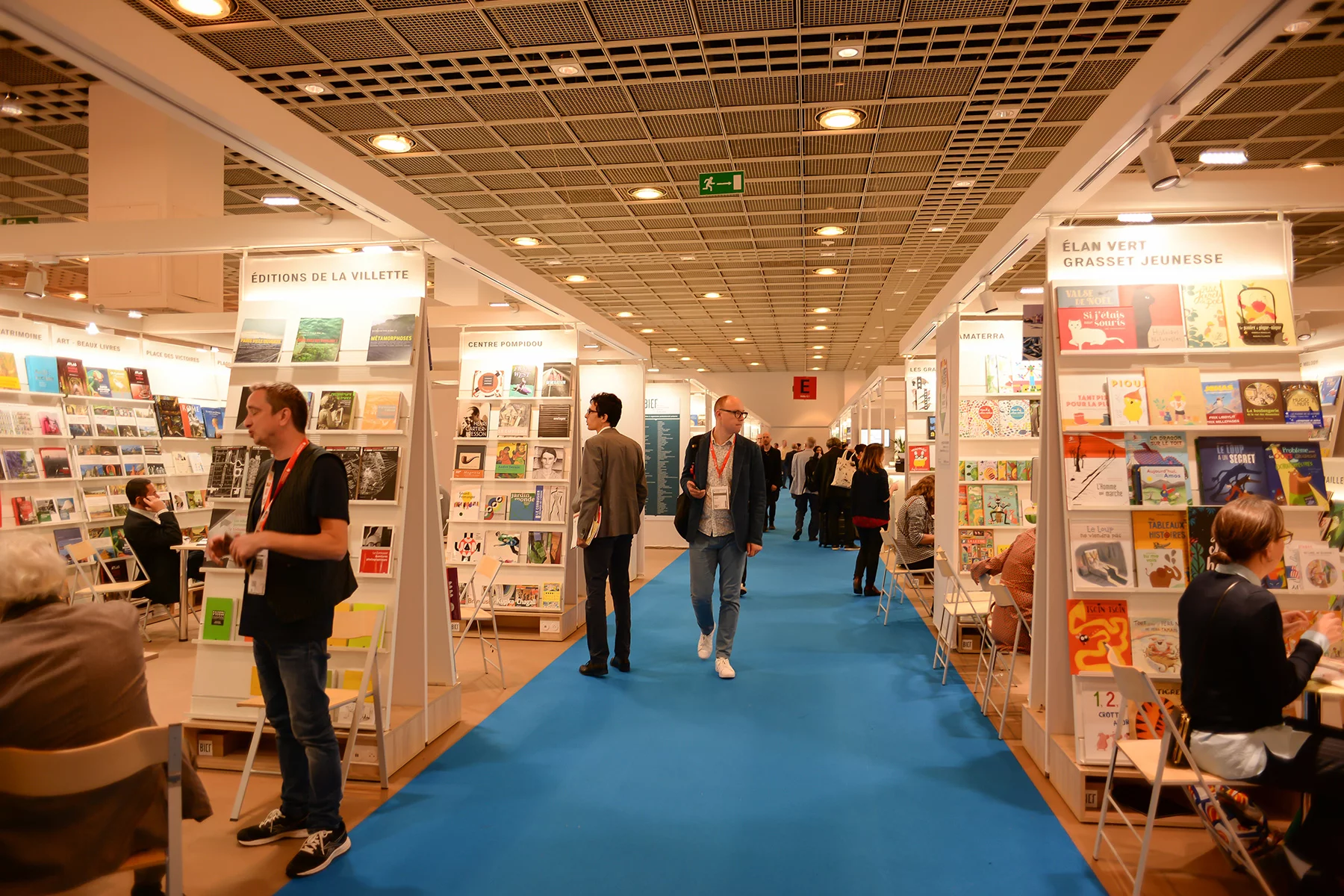 A man walks between displays of books at Frankfurt book fair. People on either side are talking to each other or looking at displays.