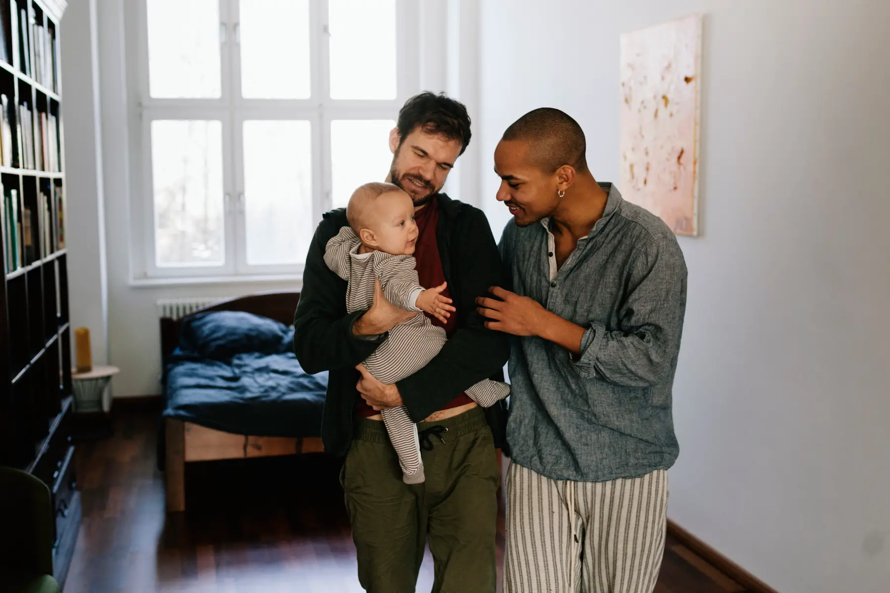 two men wearing pajamas and holding a young baby in their bedroom