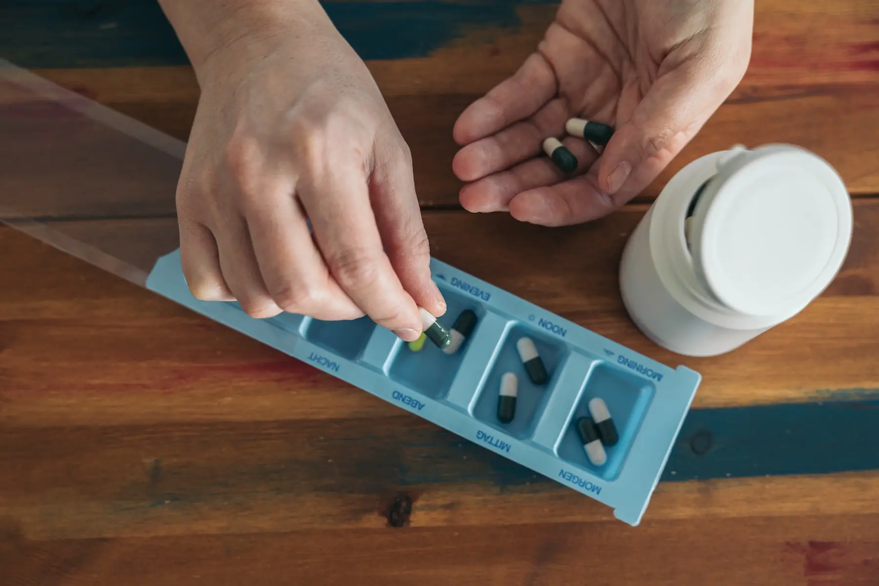A person is putting their daily medication into a pill organizer, labeled with times of day in English and German