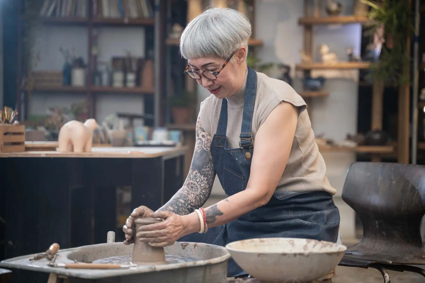 Middleaged lady with tattoos forming a clay pot with her hands in her pottery workshop.