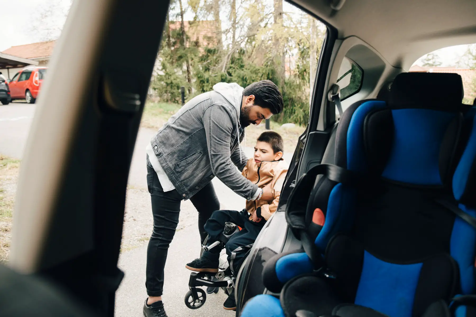 Father carrying disabled son from wheelchair seen from car.
