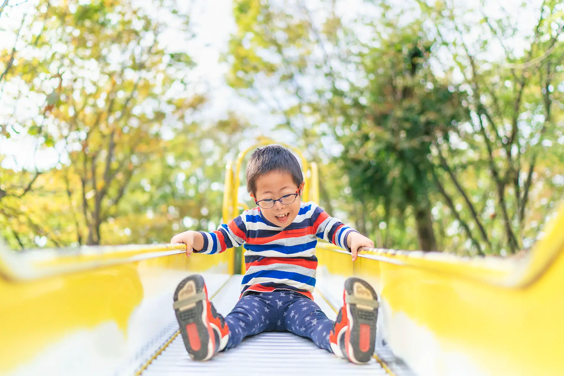 A small child with down syndrome is playing on a slide in a playground on a sunny day.