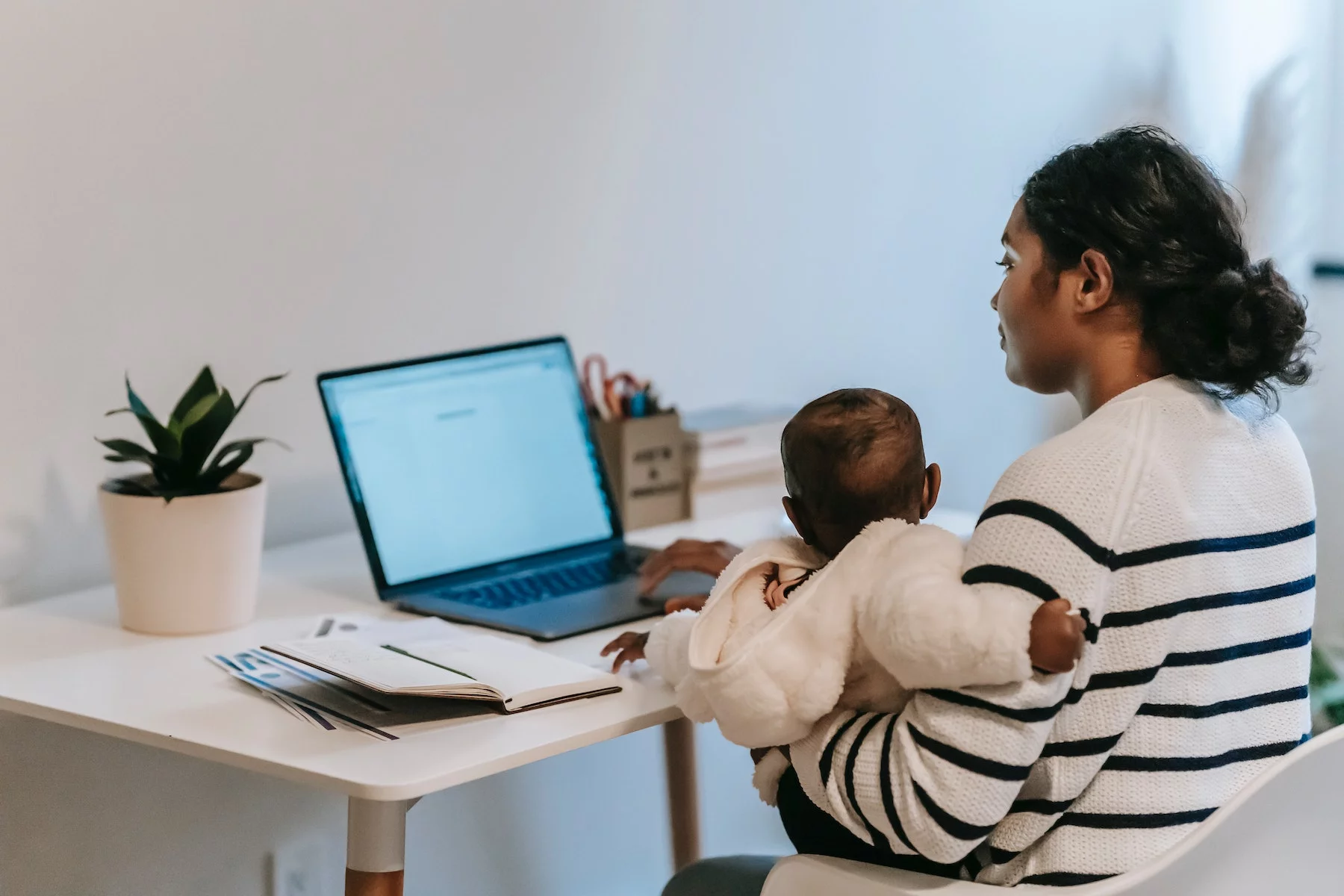 A mother sits at a desk using her laptop with one arm and holding a baby in the other.