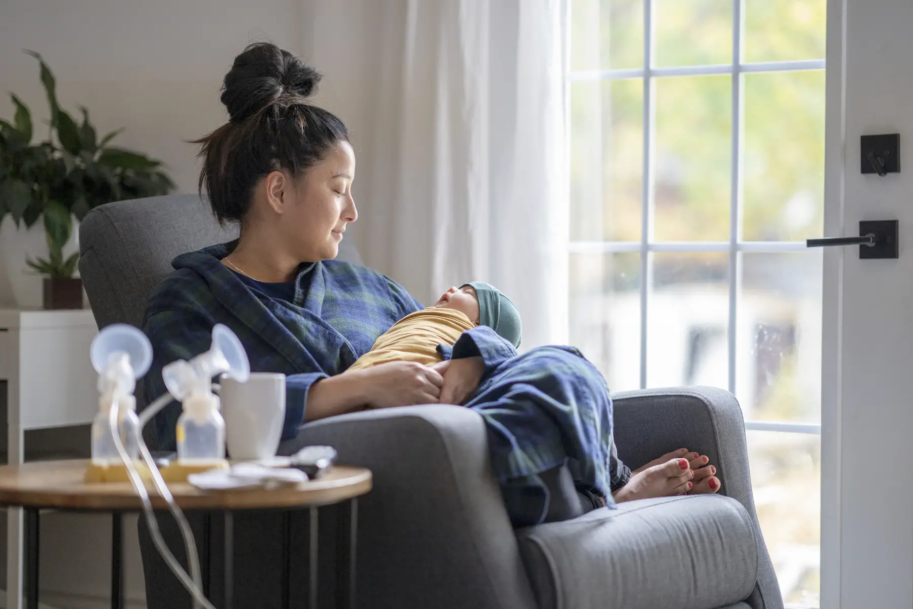 A new mother sitting on a rocking chair in her living room wearing a bathrobe, holding her newborn baby. 