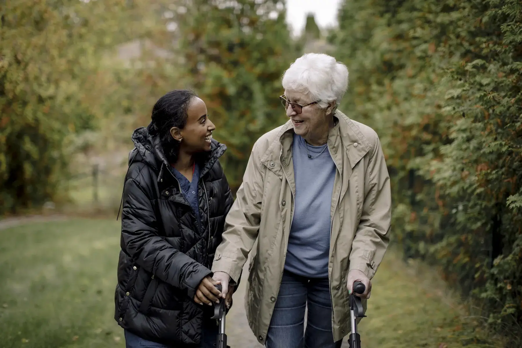 A young nurse helps an older woman walk outside through a garden as both smile