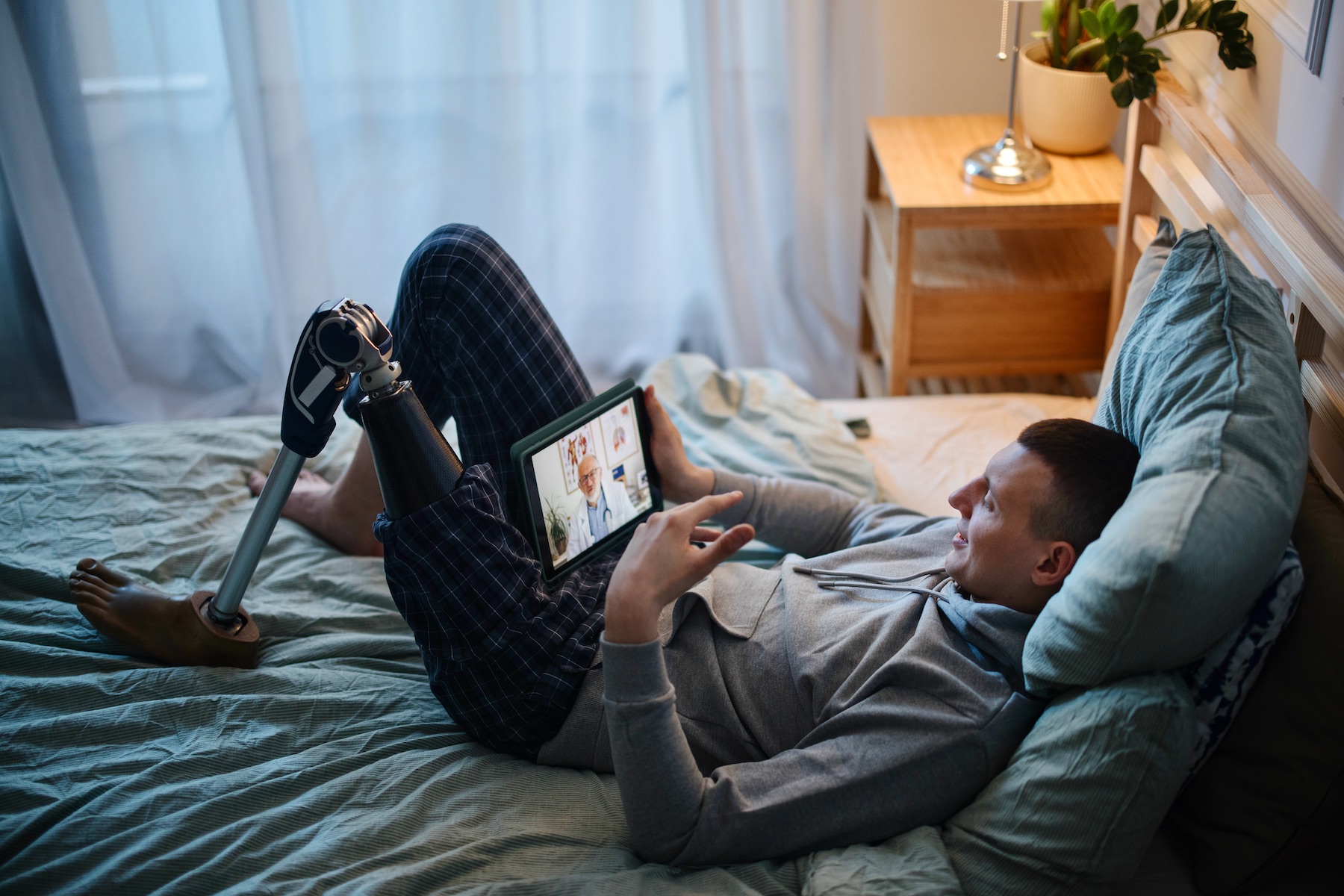 A young man leans back on a pillow in bed while chatting with his psychiatrist via video call on a tablet.
