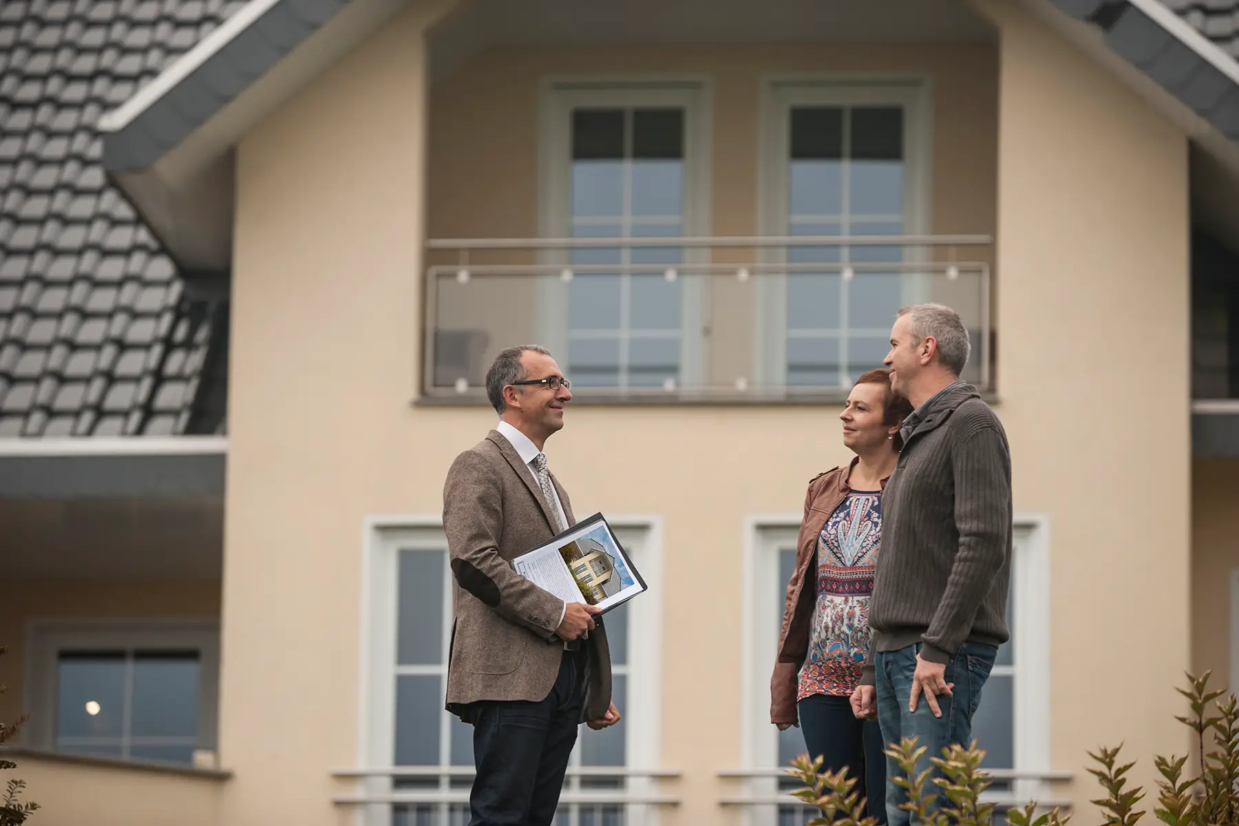 A couple stand outside a large modern house with an estate agent holding a clipboard with a picture of the house on it.
