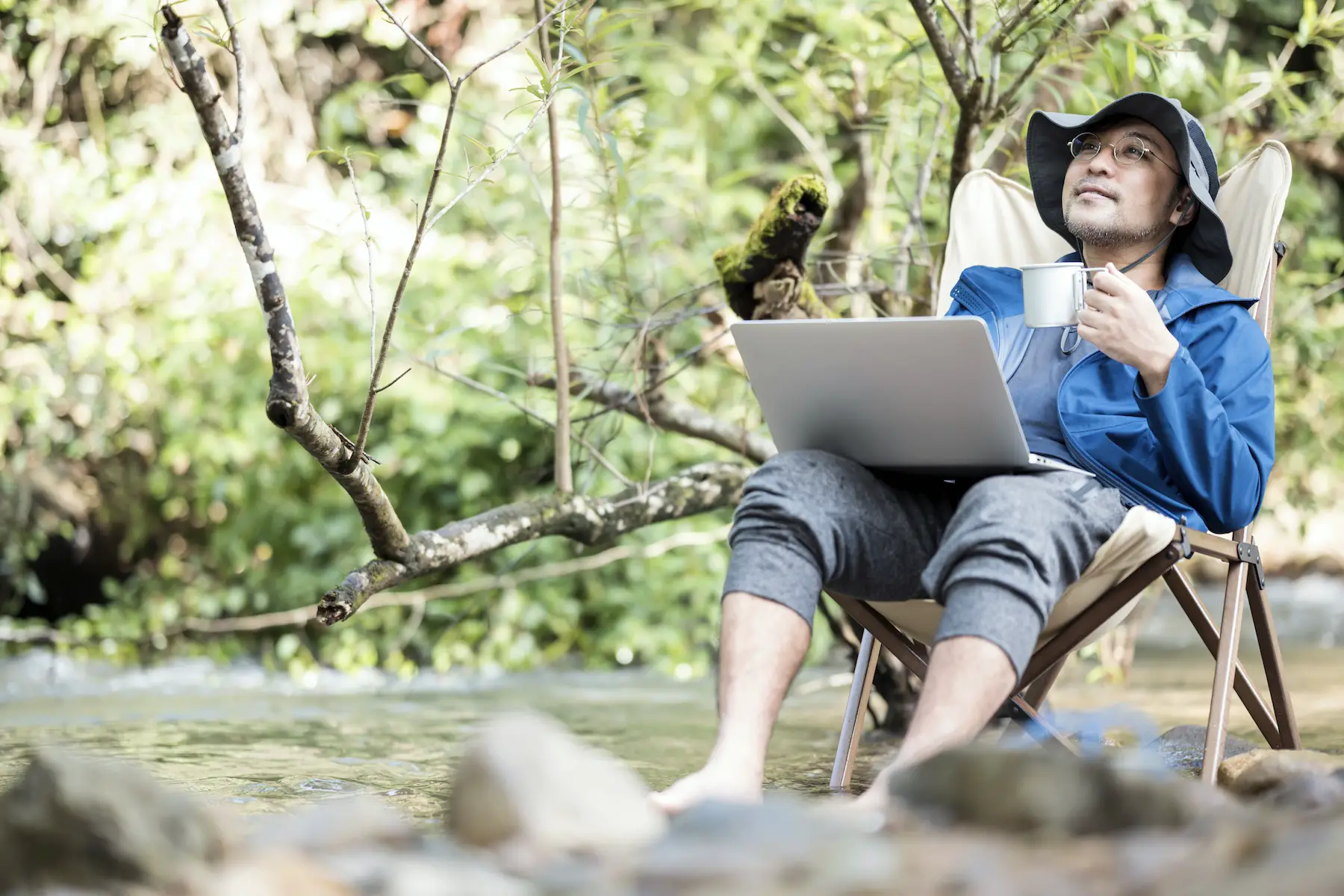 A man relaxes while working on his laptop in a forest with a mug of coffee in one hand and his feet in creek.