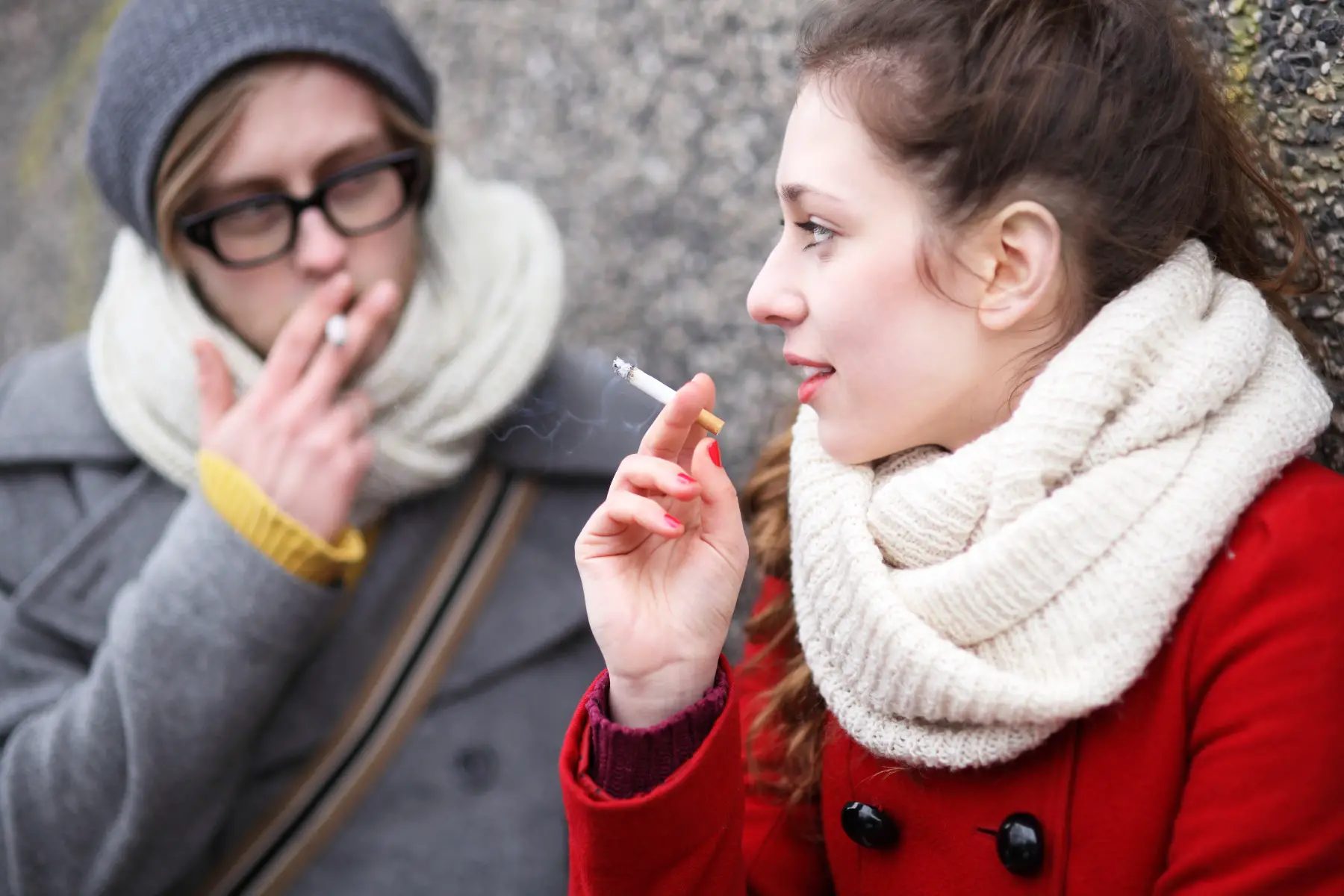 a teenager boy and girl wrapped up in winter clothing and smoking cigarettes outside while having a conversation