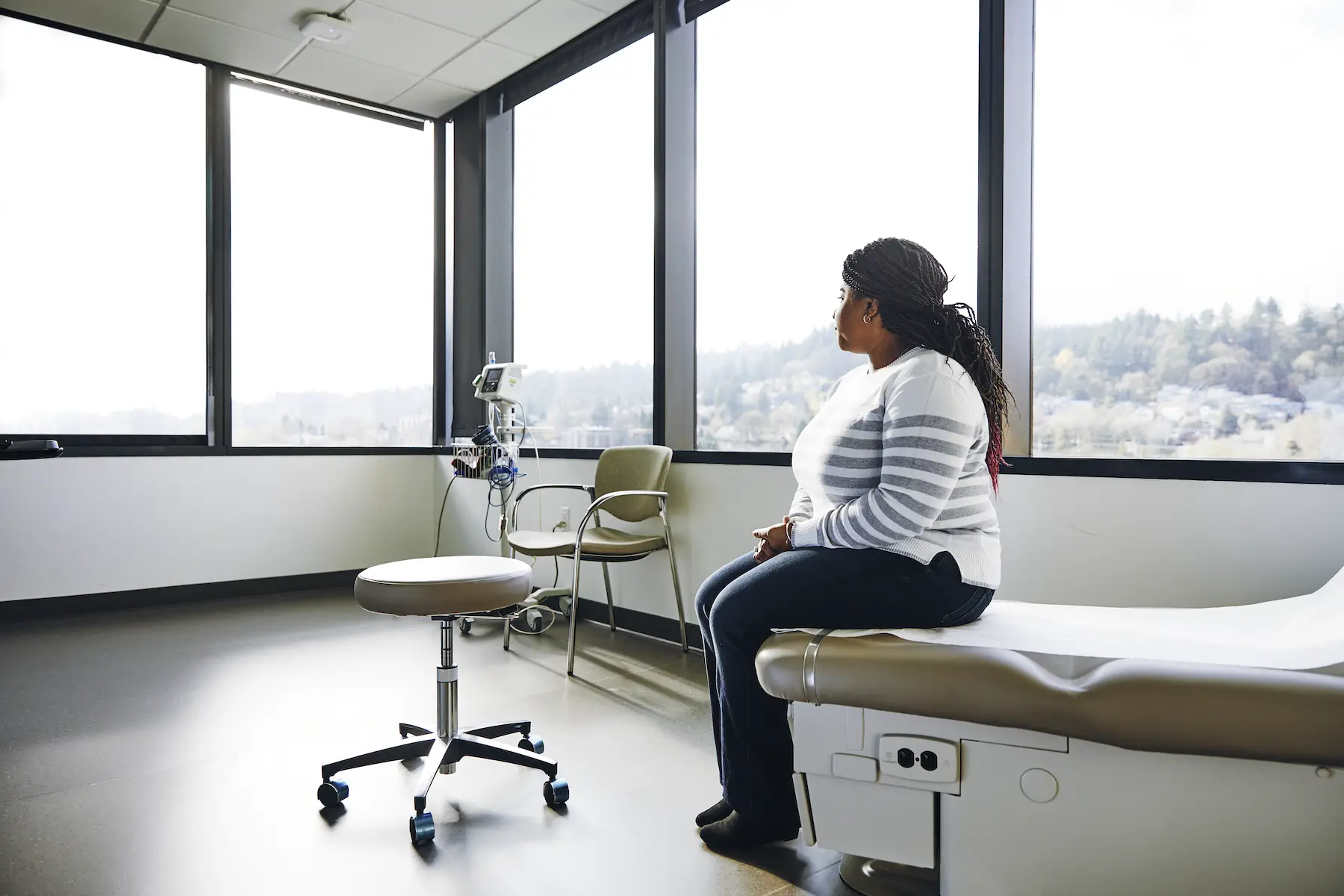 A female patient sits on the edge of an exam table in an empty doctor's office with windows overlooking a forest
