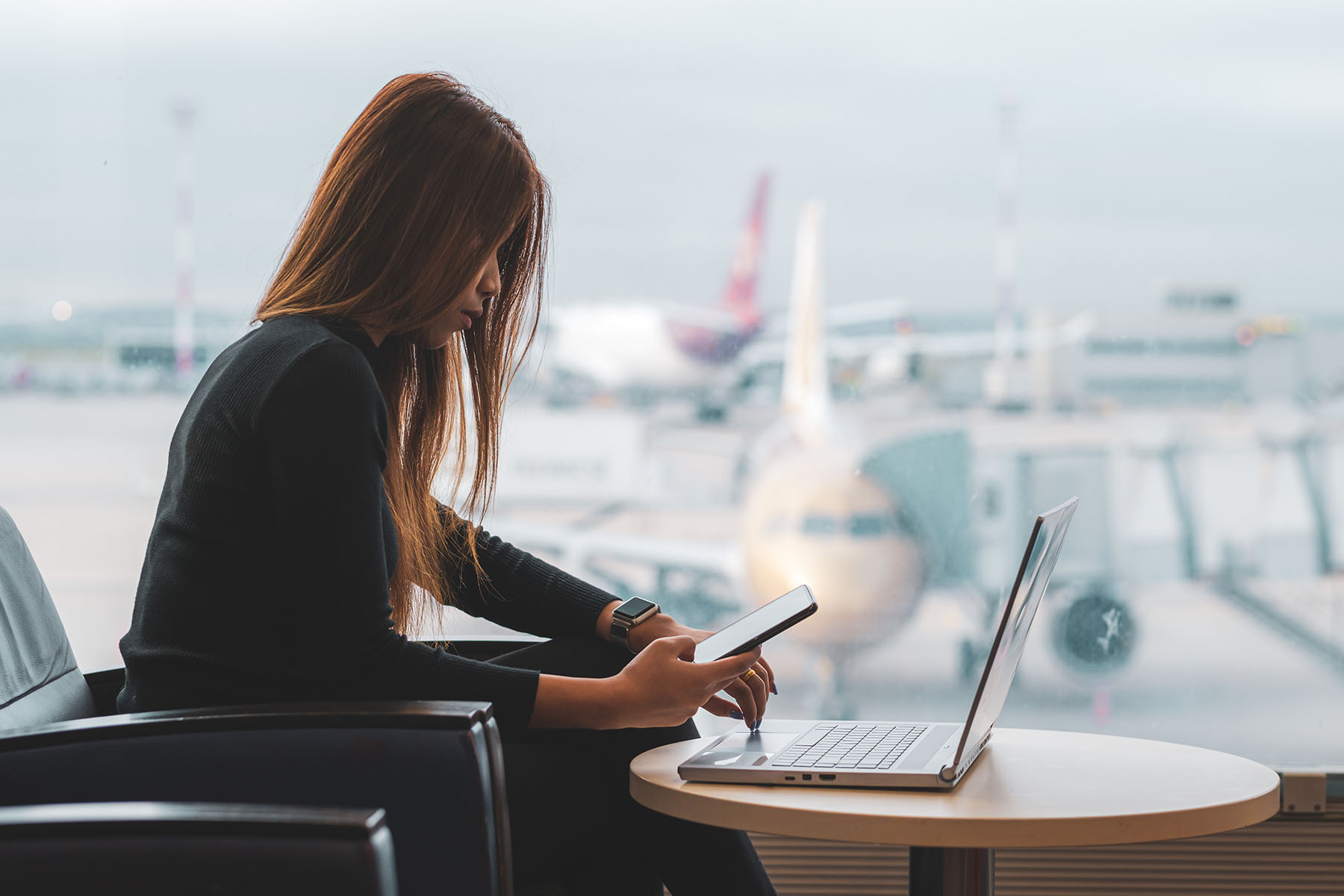 Woman working on laptop in airport hall. Planes are visible in the background.