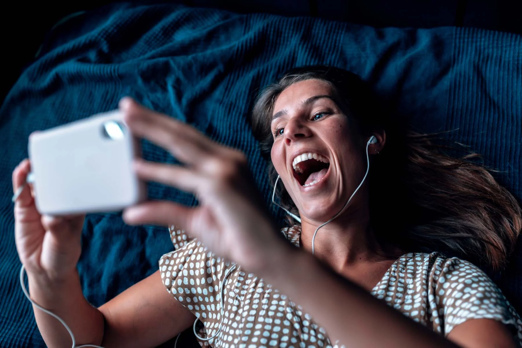 A woman looking astonished while lying down and holding a tablet with her headphones plugged into it