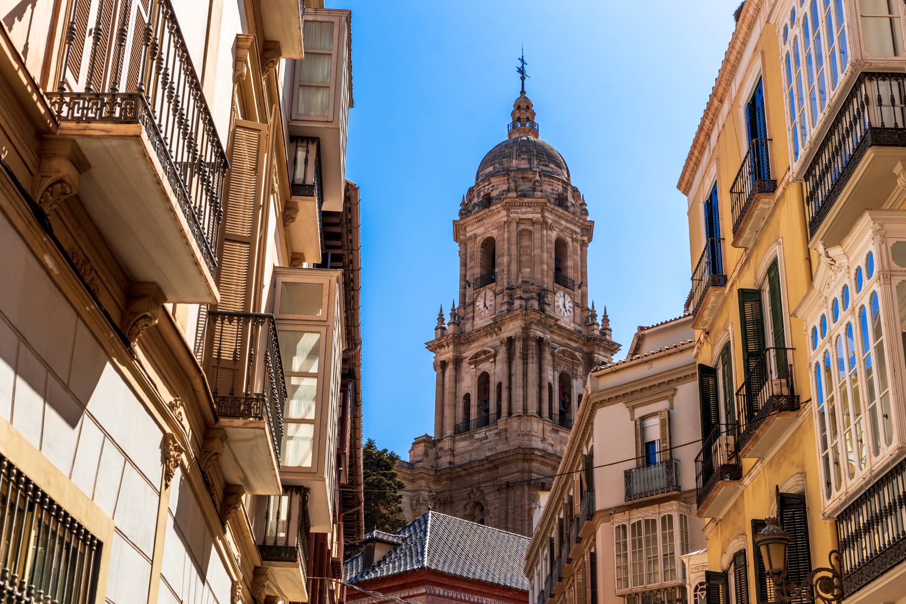 Malaga Cathedral tower seen between buildings lining a narrow street on a sunny day in Malaga, Spain