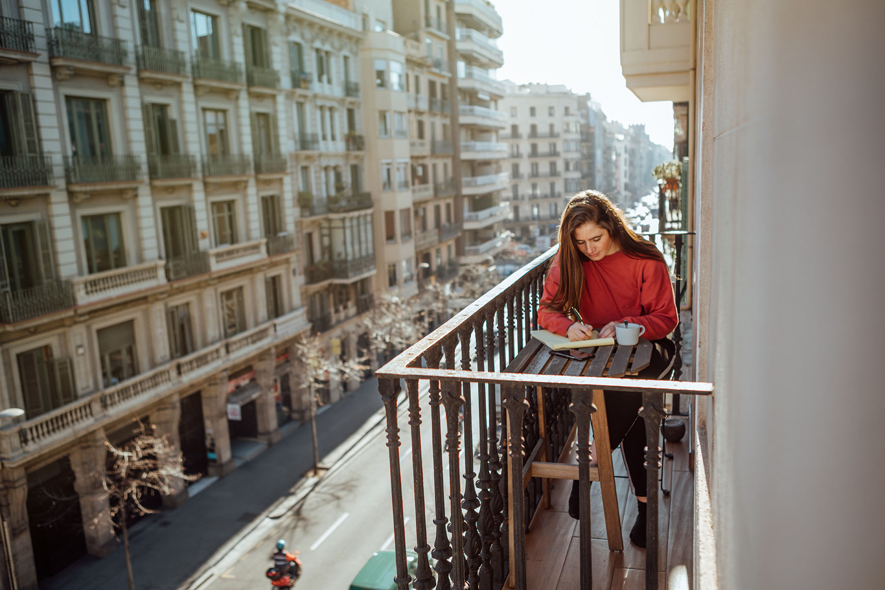 Woman sitting on a balcony, drinking out of a mug, and writing on a note pad.