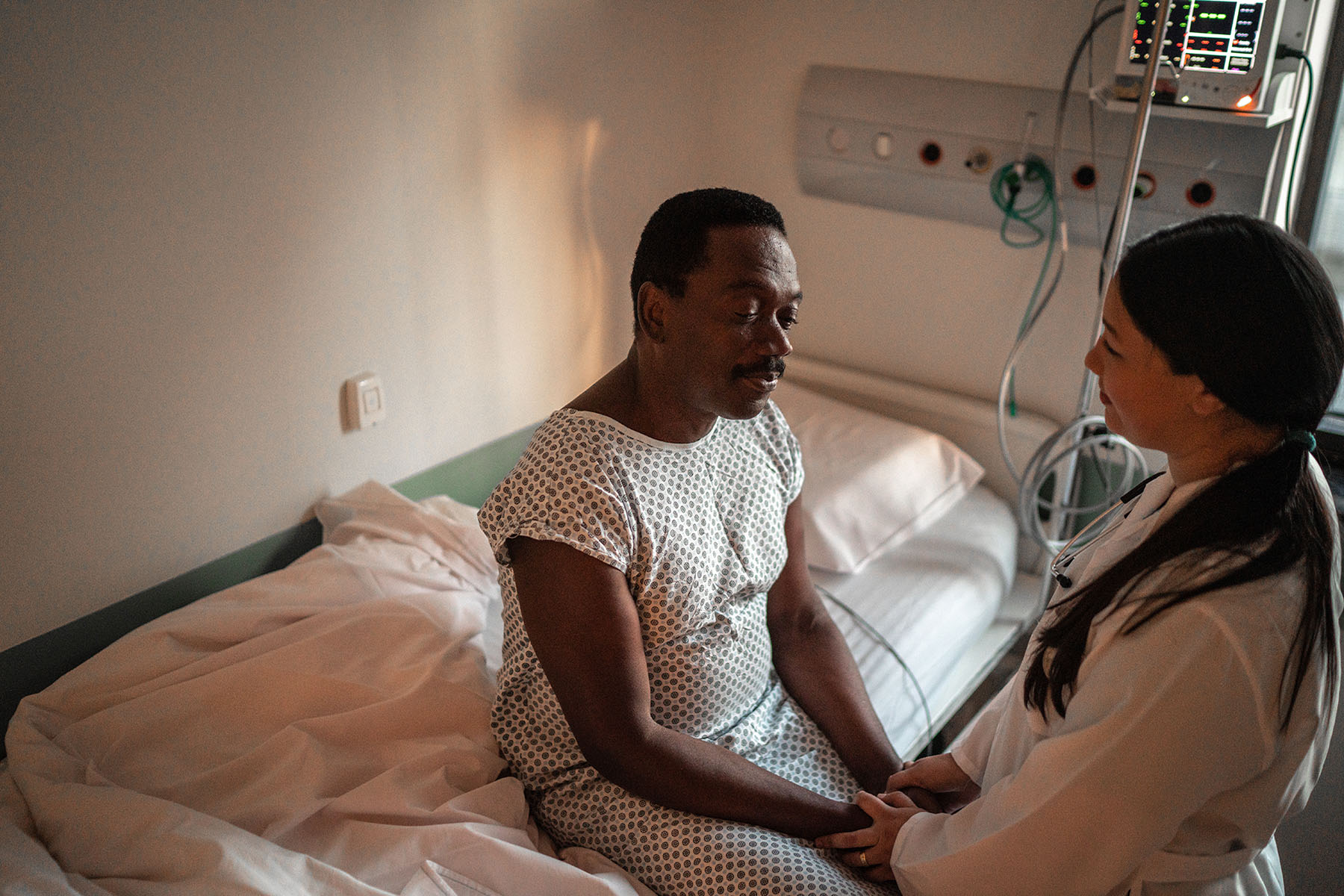 A patient sitting in a hospital gown on a bed while the doctor holds his hands